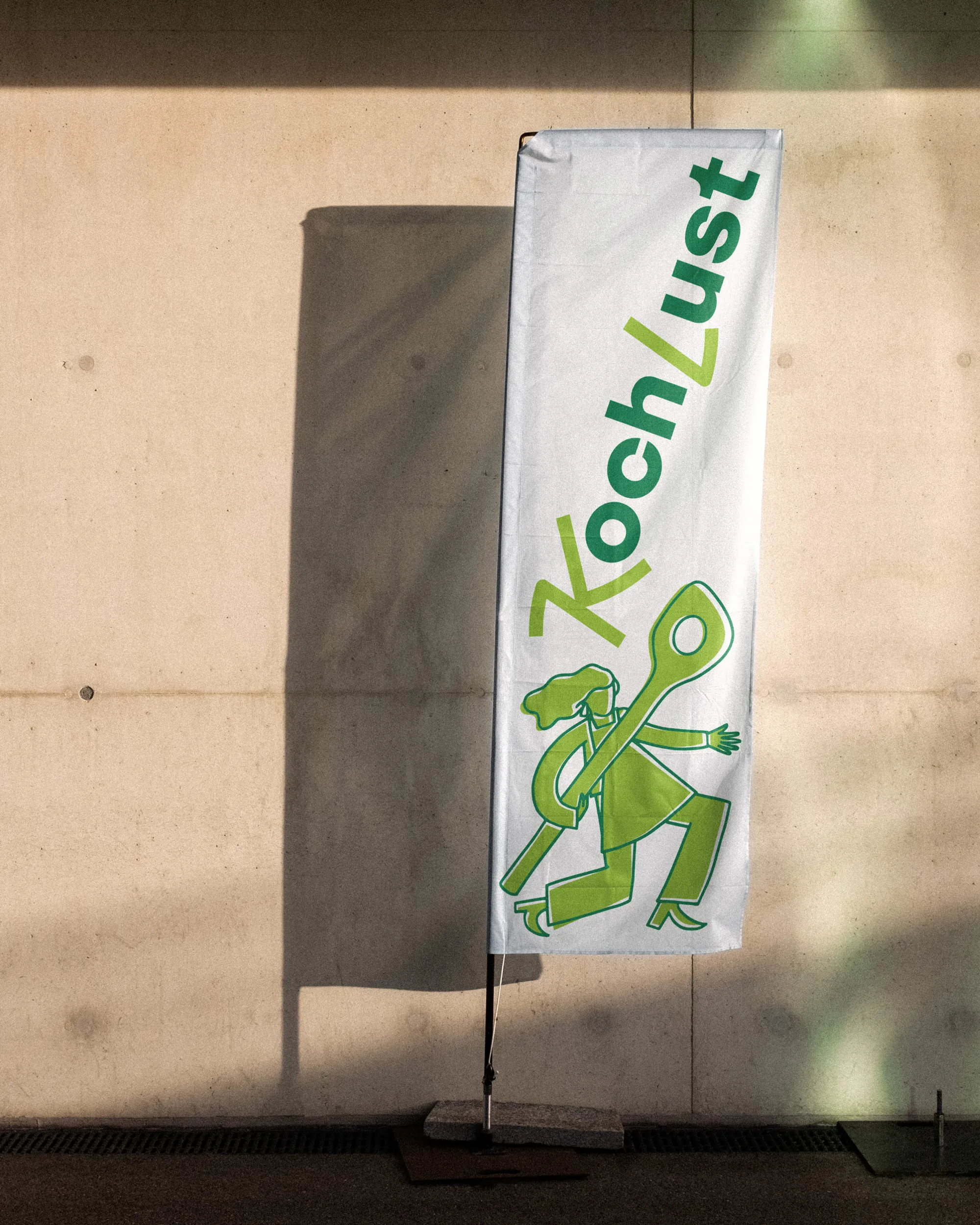 KochLust Fahne mit Logo und grüner Illustrationsfigur vor Betonwand