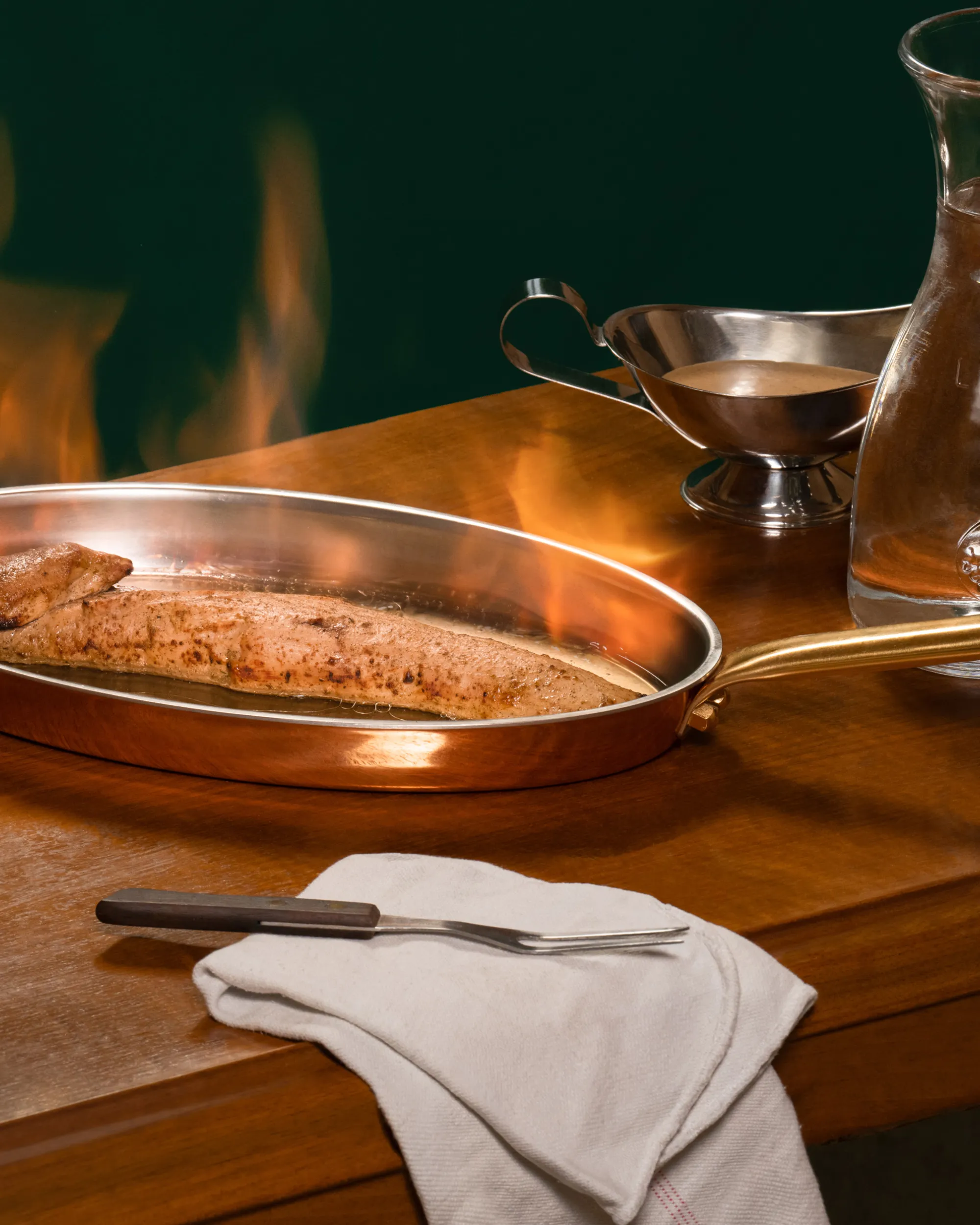 Seared fish fillet cooking in a copper pan with flames on a wooden table, accompanied by a fork on a white cloth and a sauce boat with gravy.