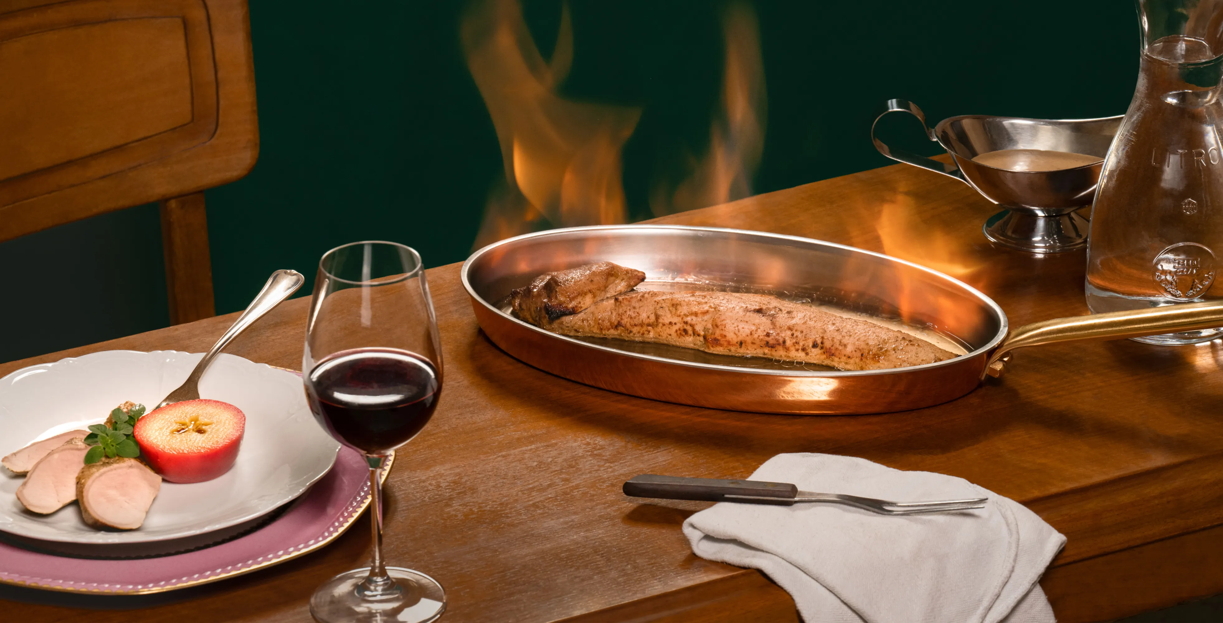 Copper pan with flaming cooked fish on wooden table next to a glass of red wine and a plate with sliced meat and half an apple.