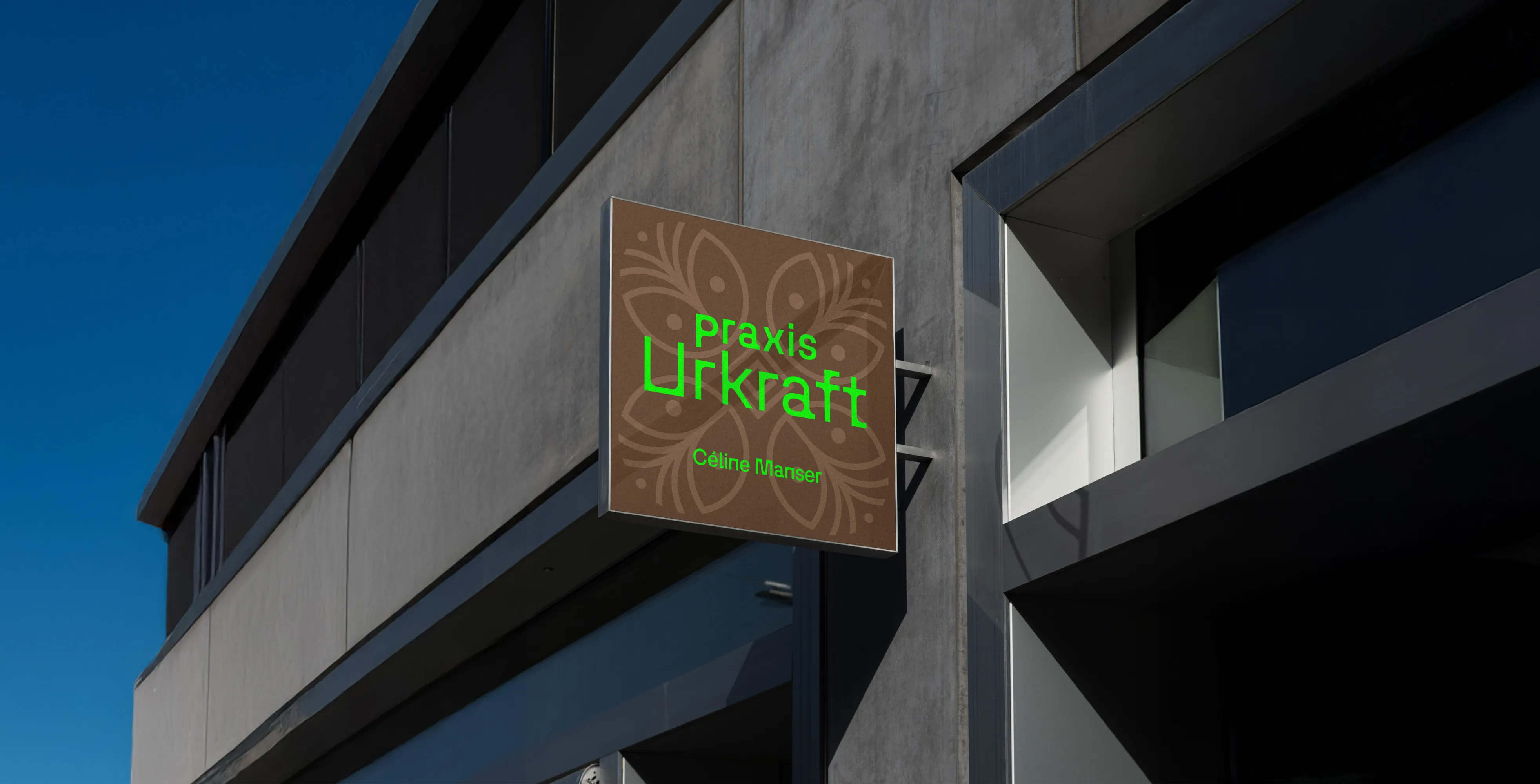 Leuchtschild der Praxis Urkraft von Céline Manser an einer Gebäudefassade, mit braunem Hintergrund, floralem Muster und grün leuchtender Schrift.