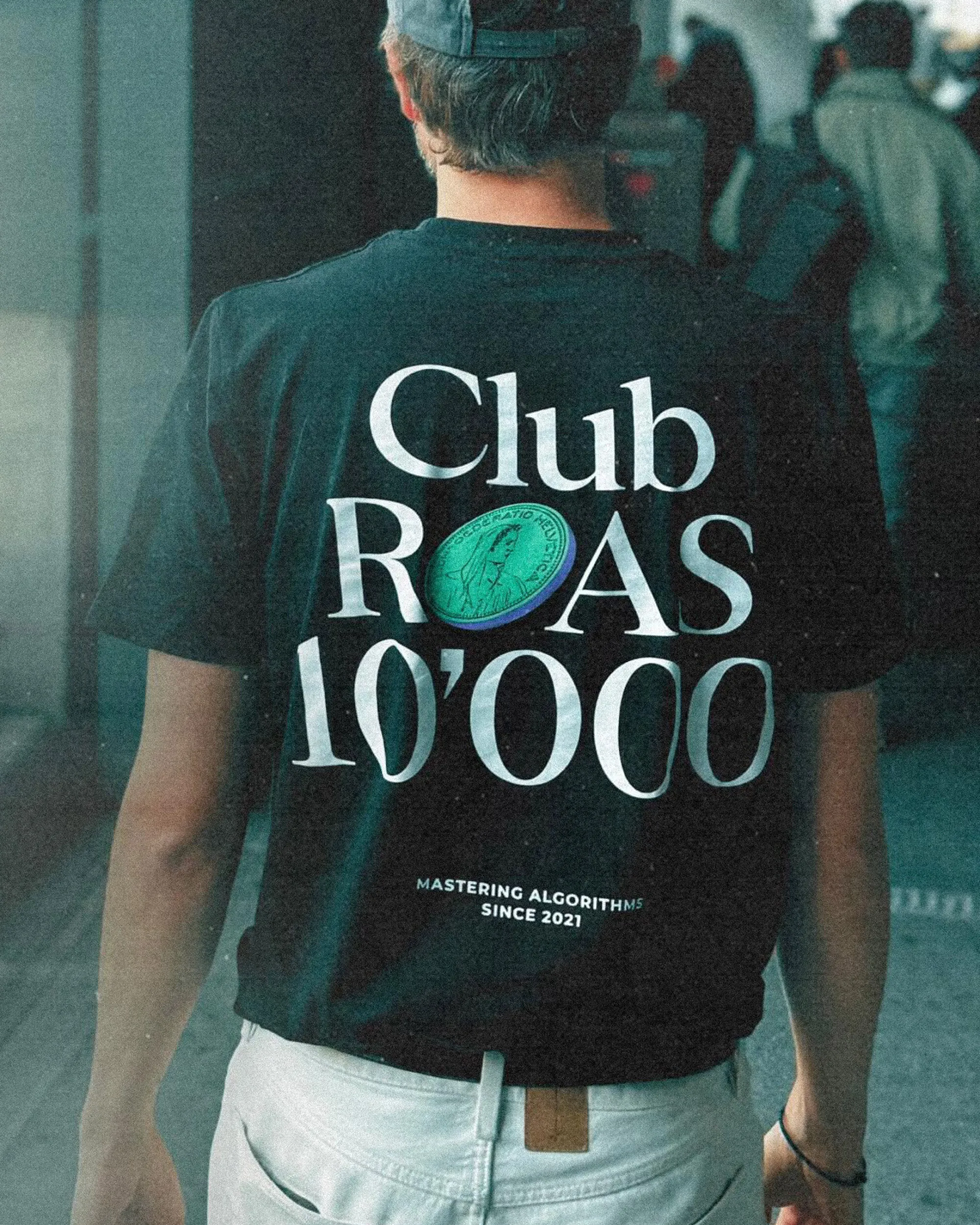 Person wearing a black t-shirt with white text 'Club ROAS 10,000' and smaller text 'Mastering Algorithms Since 2021' on the back.