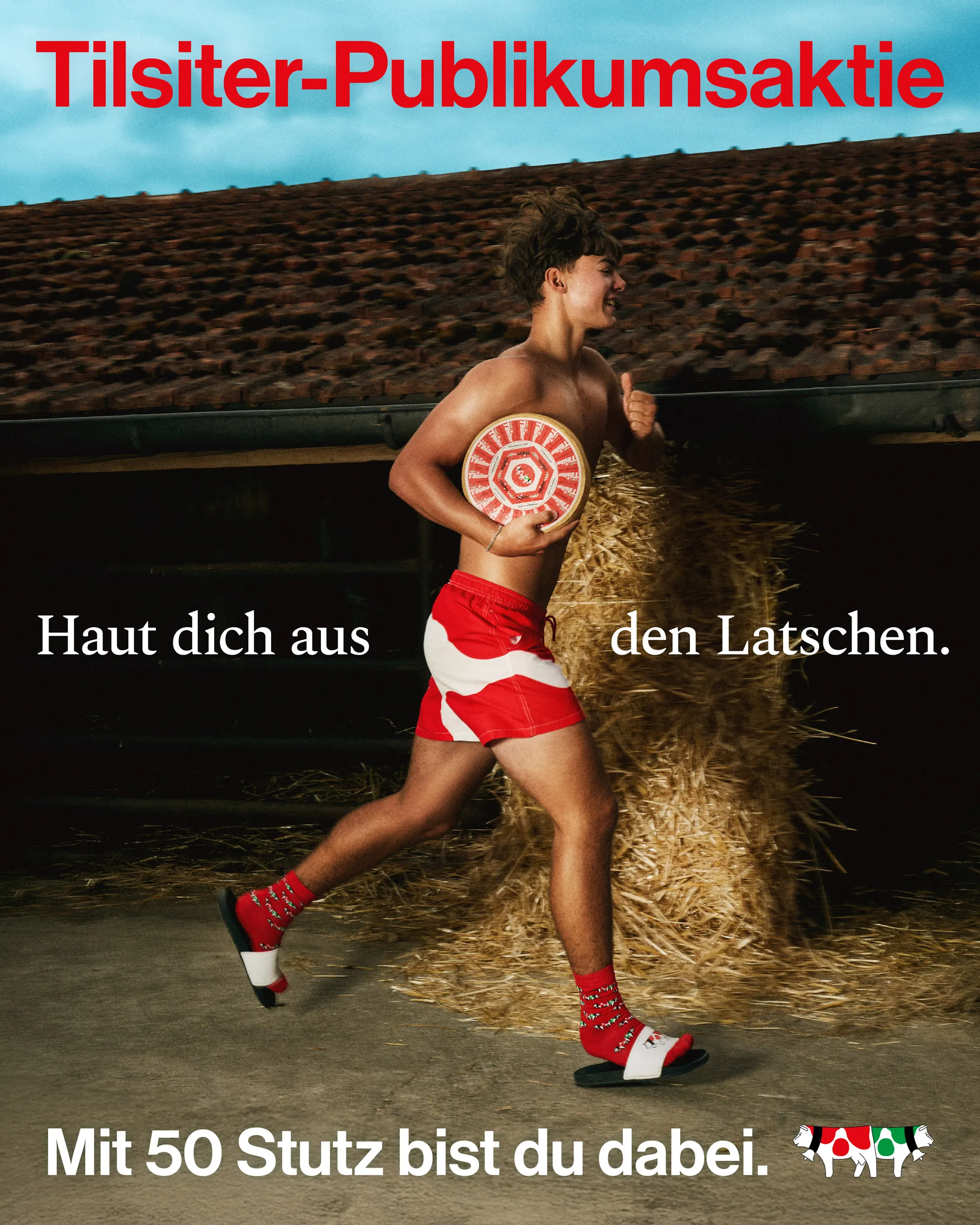 Young shirtless man running indoors wearing red shorts, matching socks, and slides, holding a Tilsiter cheese wheel and giving a thumbs-up.