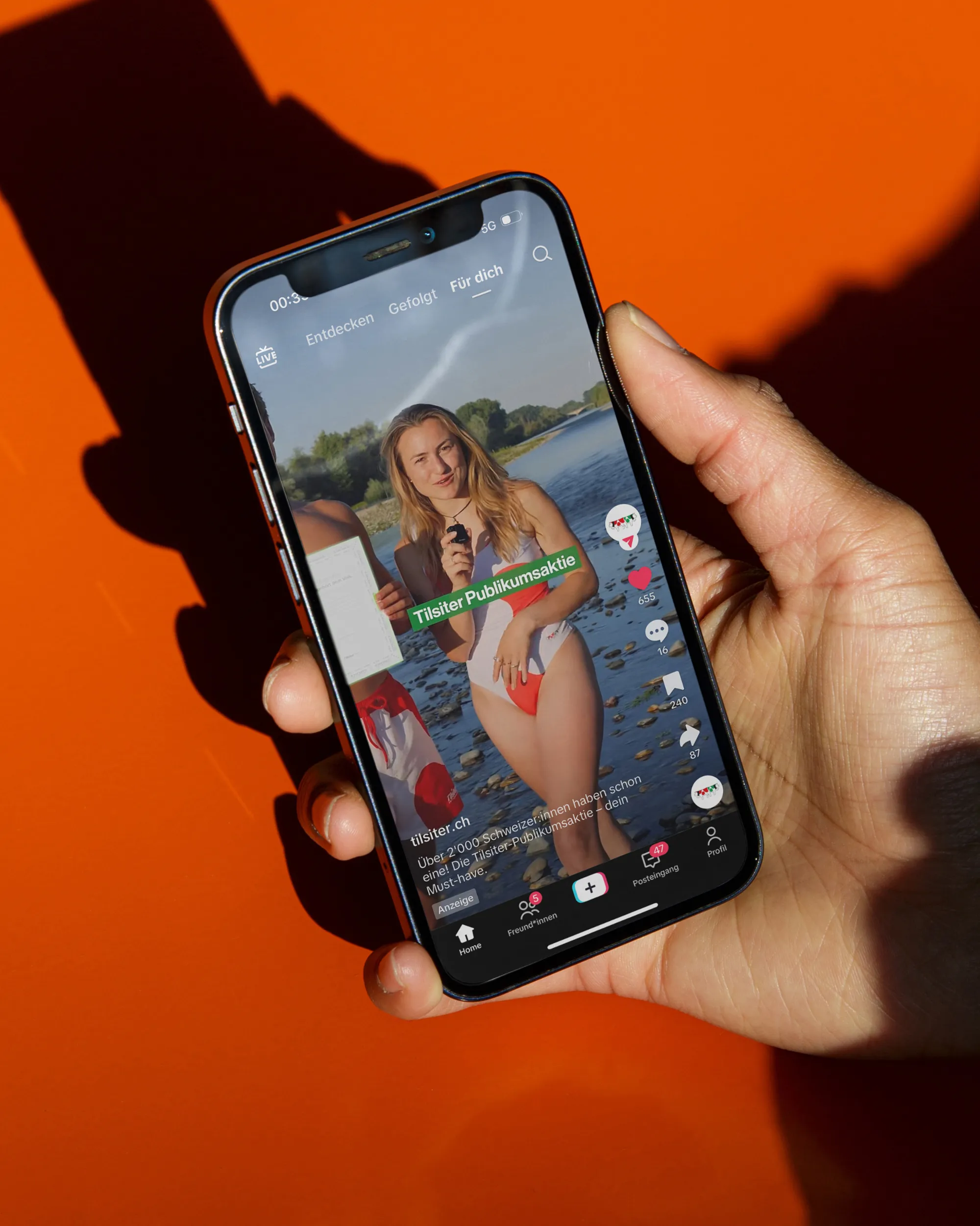 Hand hält ein Smartphone mit laufendem Instagram-Video. Auf dem Bildschirm steht eine Frau im roten Badeanzug an einem Flussufer. Im Video eingeblendeter Text: „Tilsiter Publikumsaktie“. Darstellung einer Instagram-Videoanzeige.