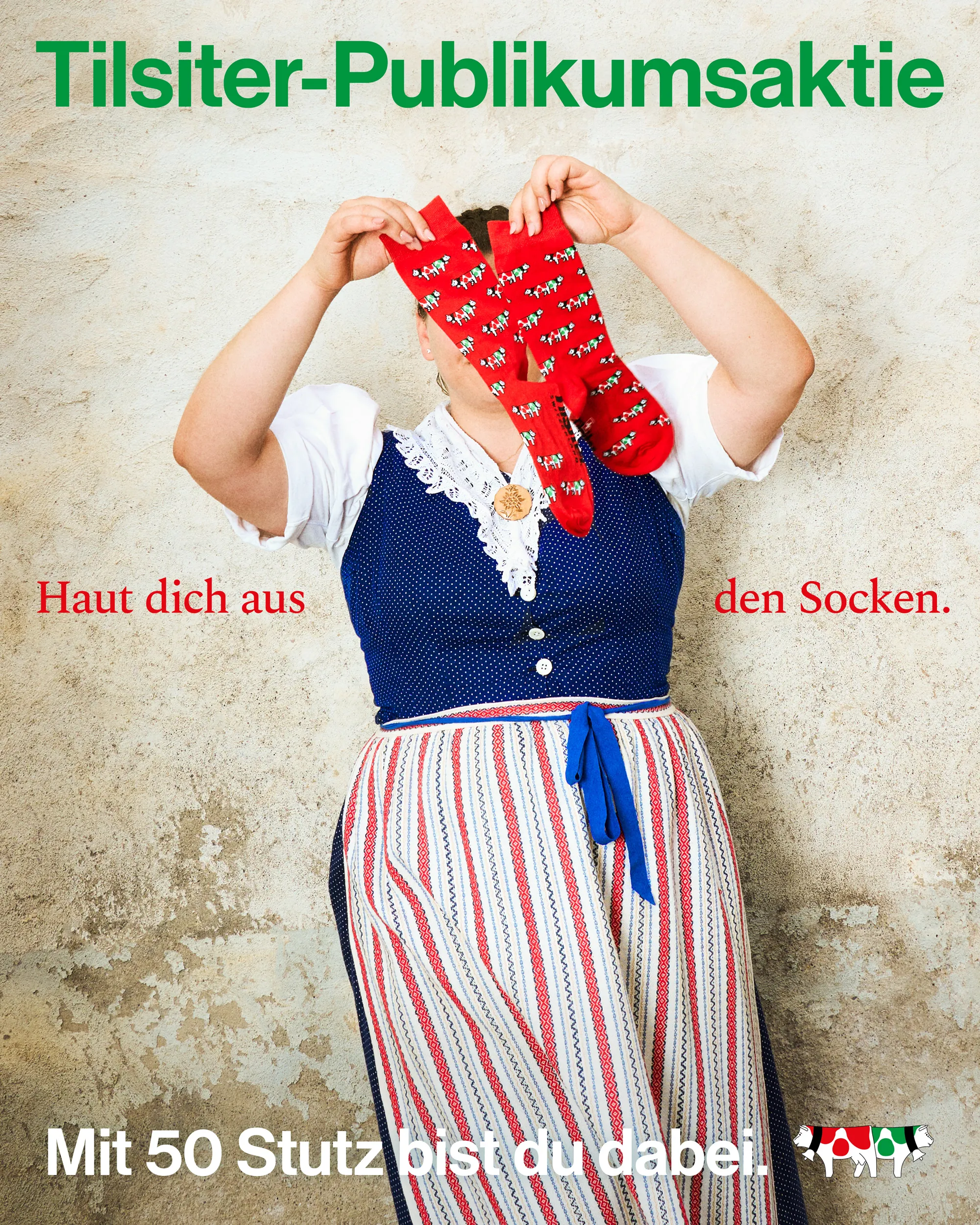 Person in traditional dress holding up two red socks with cow patterns against a textured wall background.