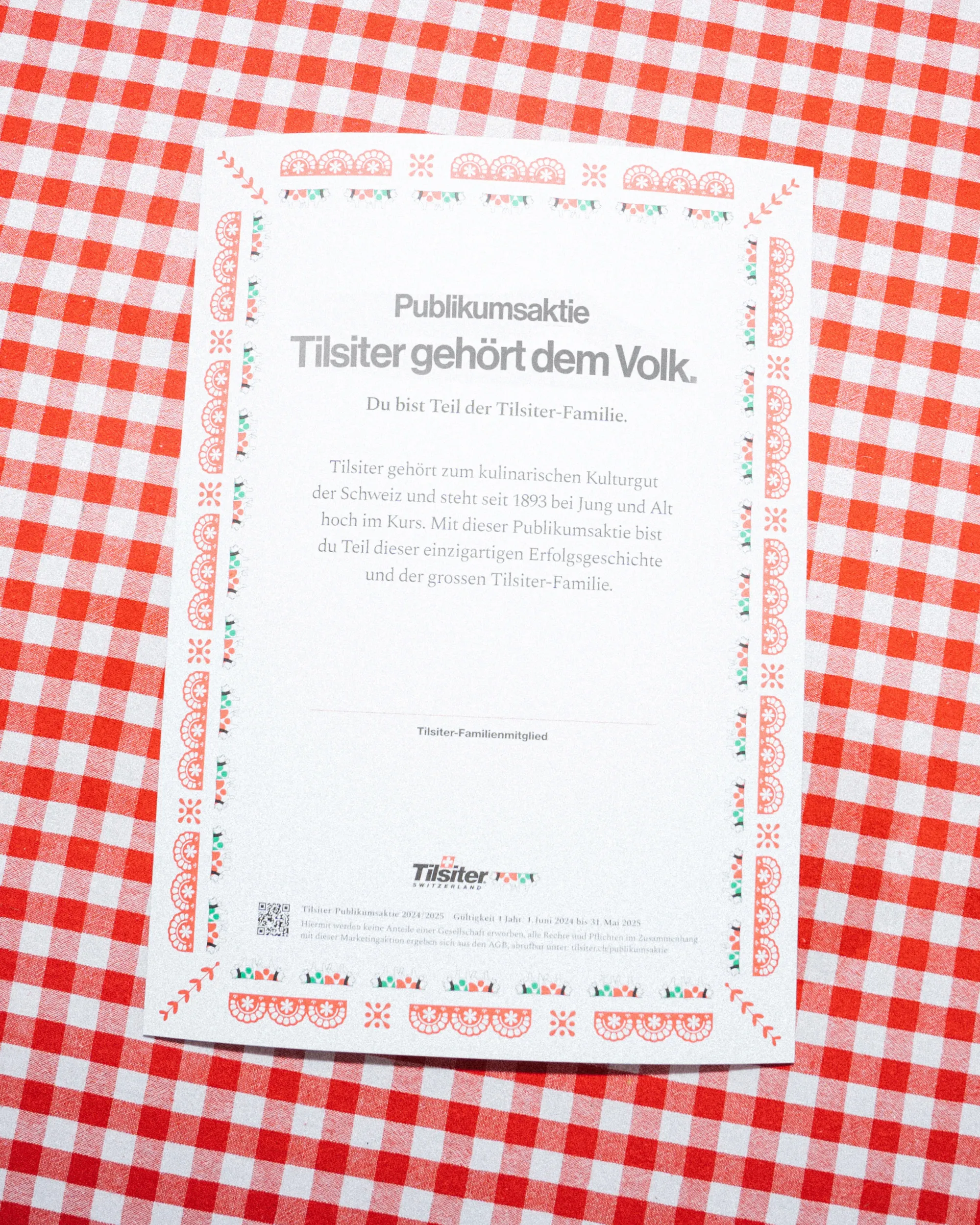 Gedruckte Schmuckaktie der Tilsiter-Publikumsaktion ‹Tilsiter gehört dem Volk› auf rot-weiss kariertem Tischtuch, mit ornamentalem Rahmen und Tilsiter-Switzerland-Logo.“