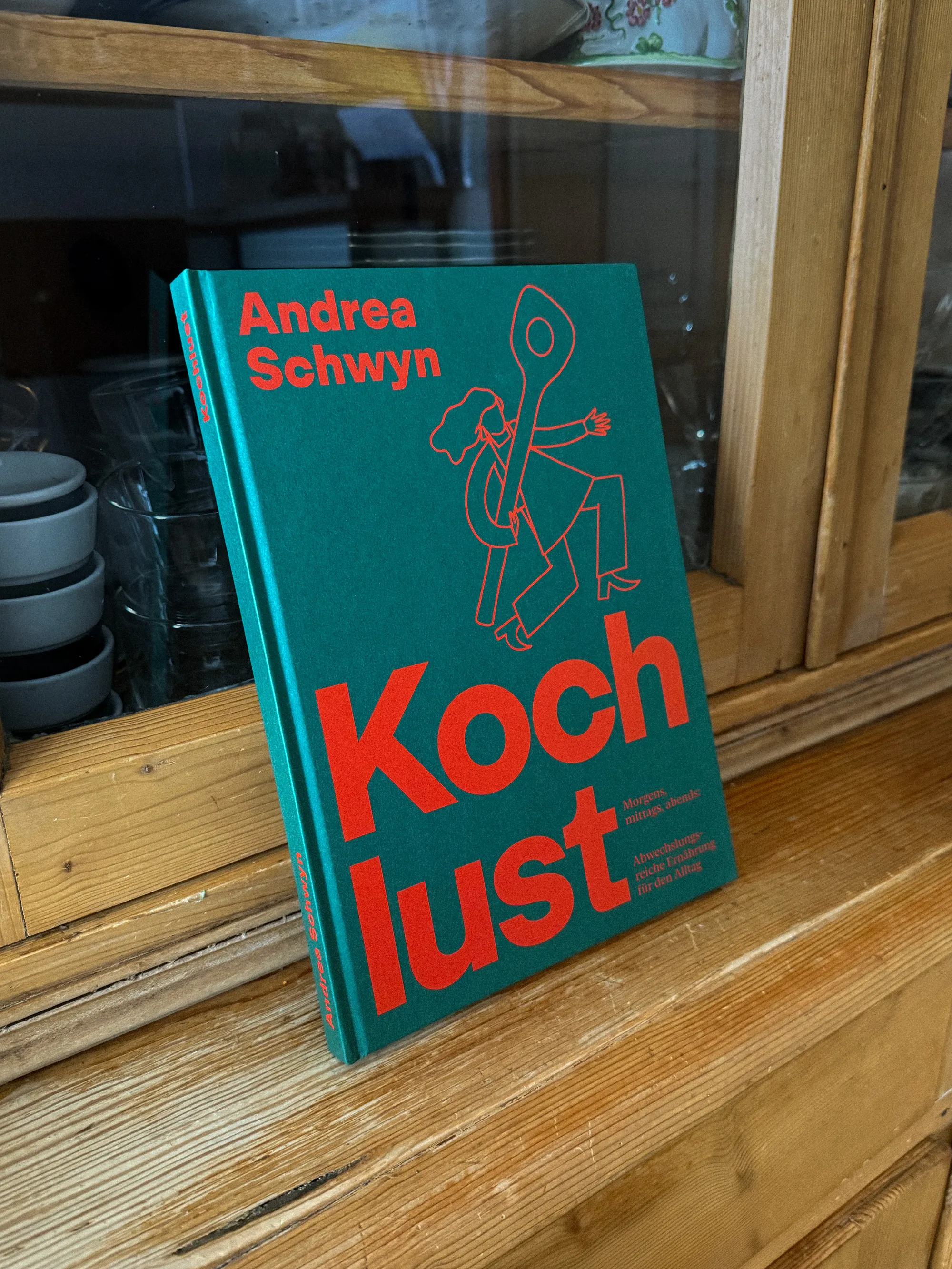 Cover des Kochbuchs Kochlust von Andrea Schwyn in Grün mit roter Typografie, aufgestellt auf einer Holzkommode vor einem Küchenschrank.
