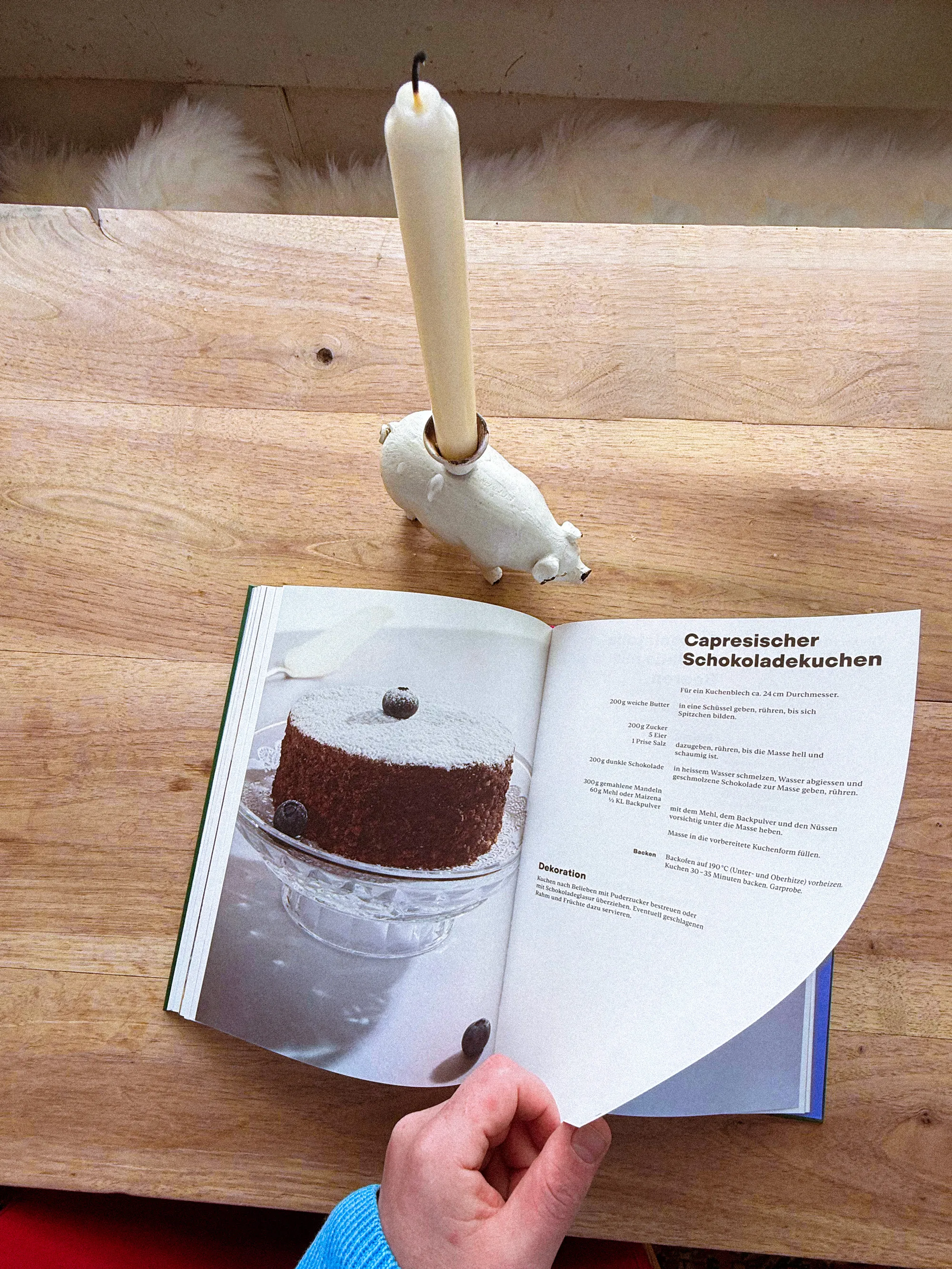 Geöffnetes Kochbuch mit Rezeptseite „Capresischer Schokoladekuchen“ und Bild eines mit Puderzucker bestäubten Kuchens; daneben steht eine weisse Kerze auf einem Holztisch.