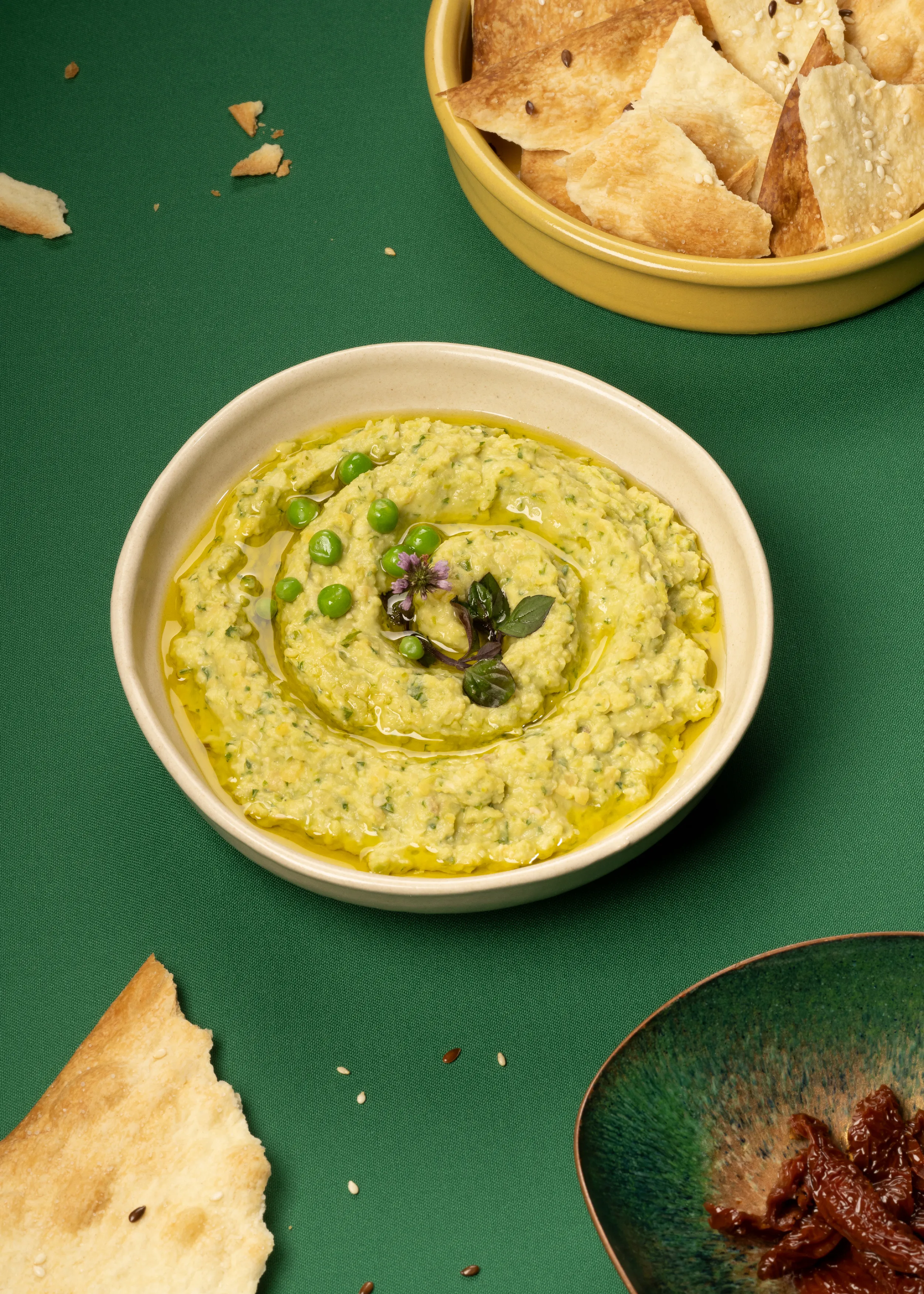 Cremiger Erbsen-Hummus in heller Schale, mit Olivenöl beträufelt und mit Erbsen und Kräutern garniert, serviert auf grünem Untergrund mit Brotstücken und Schale im Hintergrund.