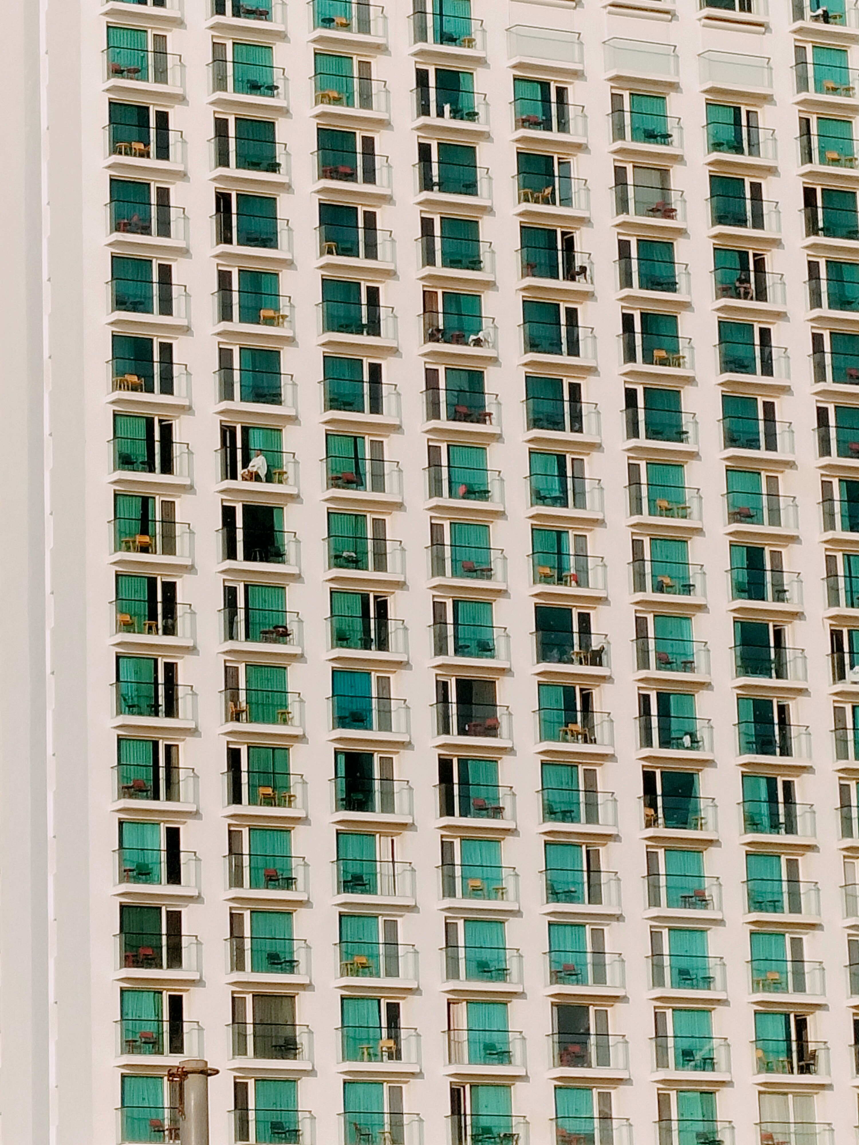 High-rise building facade with multiple balconies and teal glass doors, some with chairs and one person sitting.