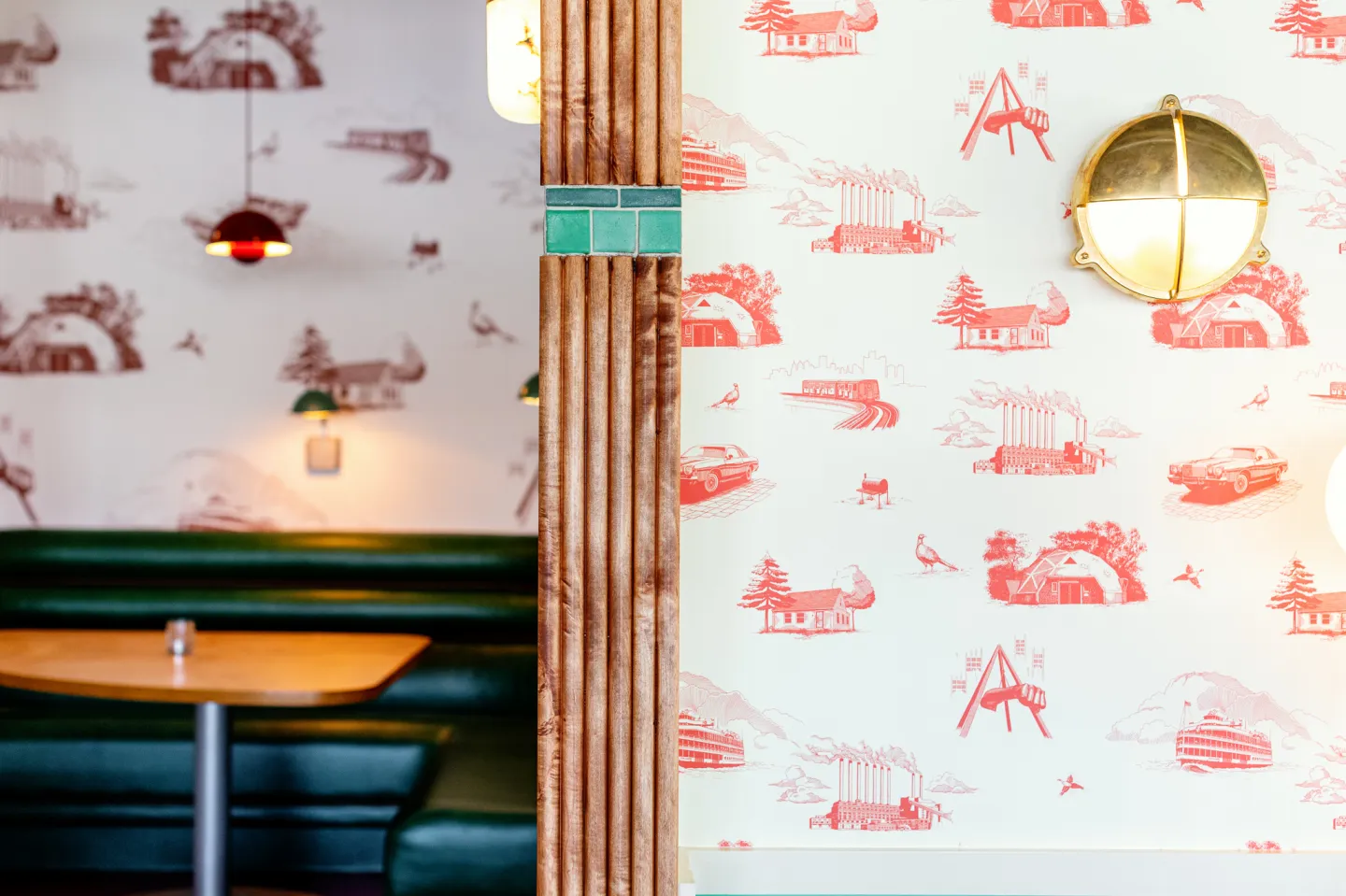 Interior view of a dining booth with green cushioned seating separated by a wooden divider, featuring walls decorated with red and white vintage-style illustrations and round light fixtures.
