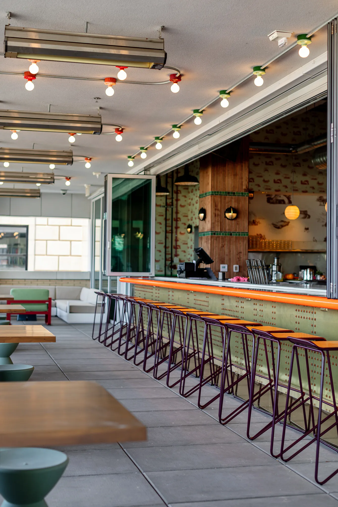 Modern bar with orange-trimmed counter and tall stools, adjacent to outdoor seating and large open windows.