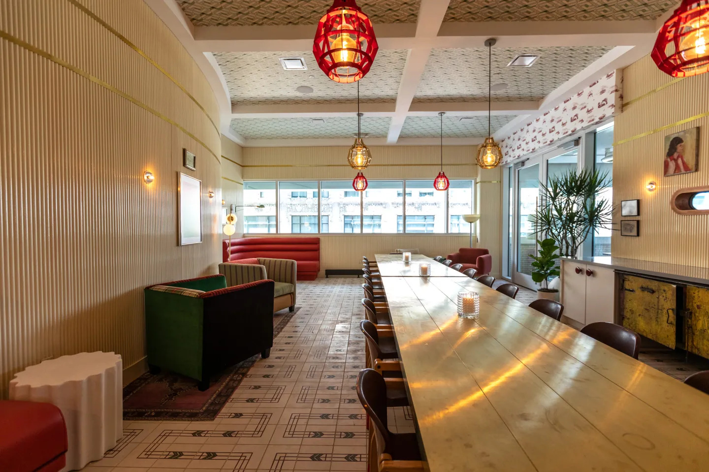 Bright, modern meeting room with a long wooden table surrounded by chairs, colorful armchairs, large windows, and hanging pendant lights.