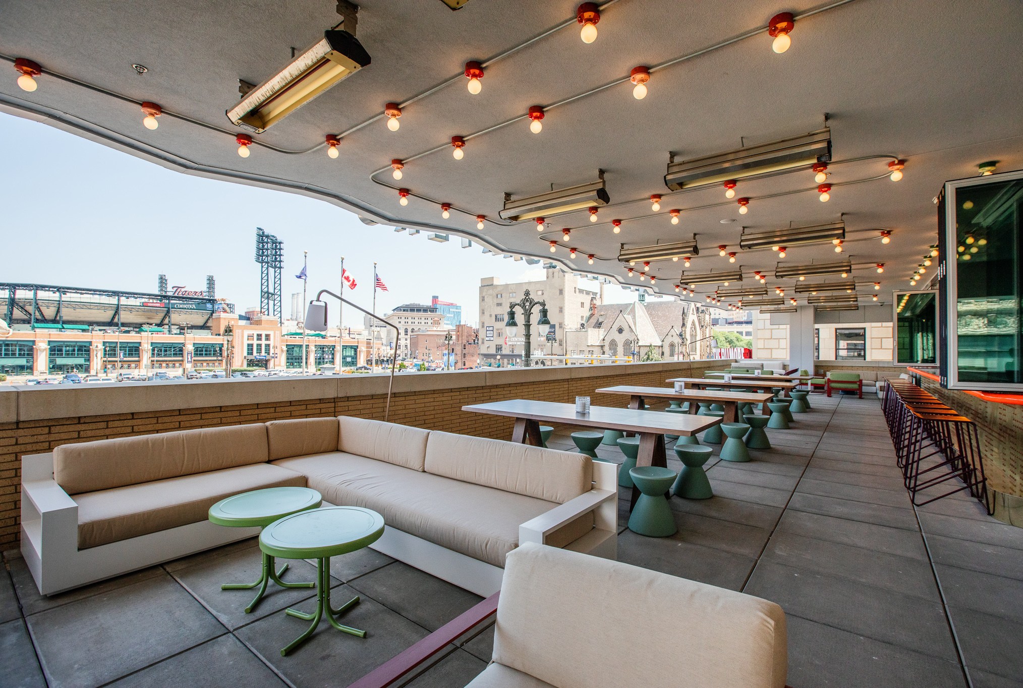 Covered outdoor lounge with beige sectional sofa, green tables and stools, picnic tables, and a view of a Comerica Park and city buildings.