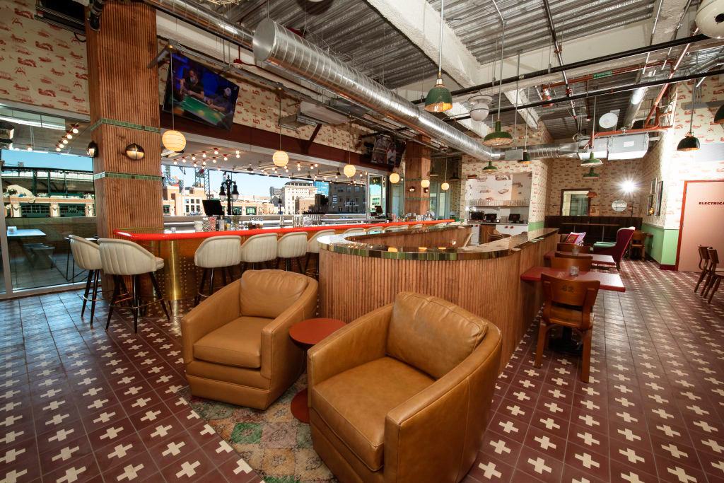 Spacious retro-style bar interior with leather armchairs, high stools at a curved wooden bar, patterned tile floor, and hanging lights.