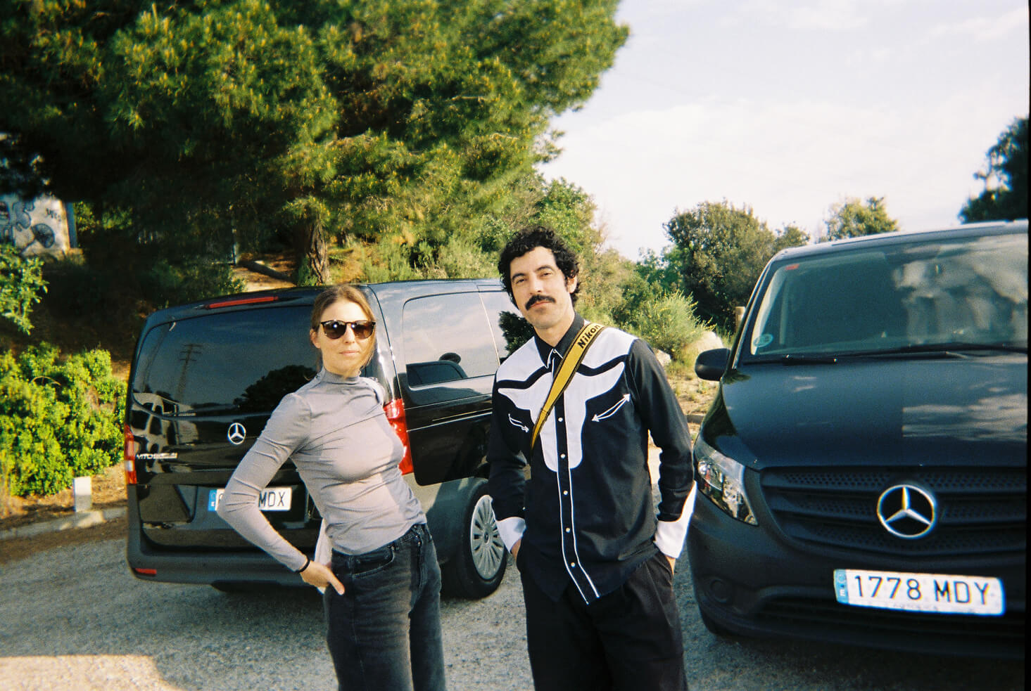 A man and a woman standing in front of two black Mercedes vans outdoors with trees in the background.