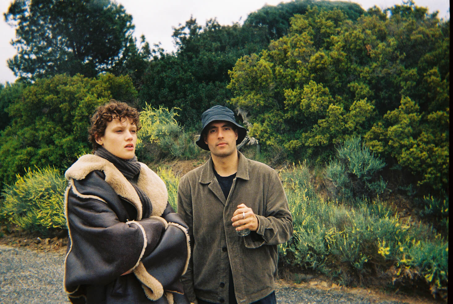 Two people outdoors with dense green bushes in the background; one wearing a large shearling coat, the other in a bucket hat holding a glass bottle.