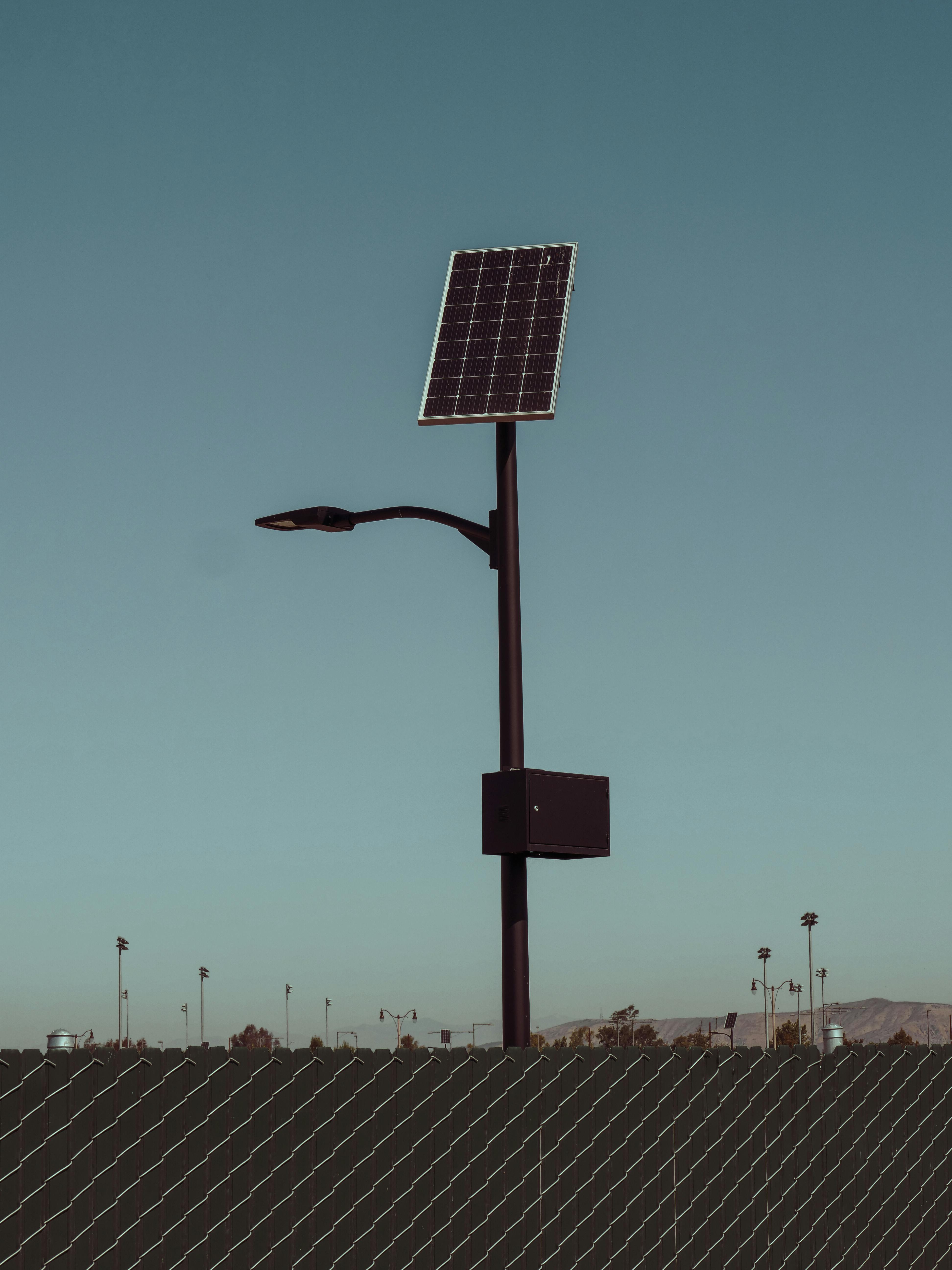 Solar-powered streetlight with a solar panel on top against a clear blue sky, behind a chain-link fence.