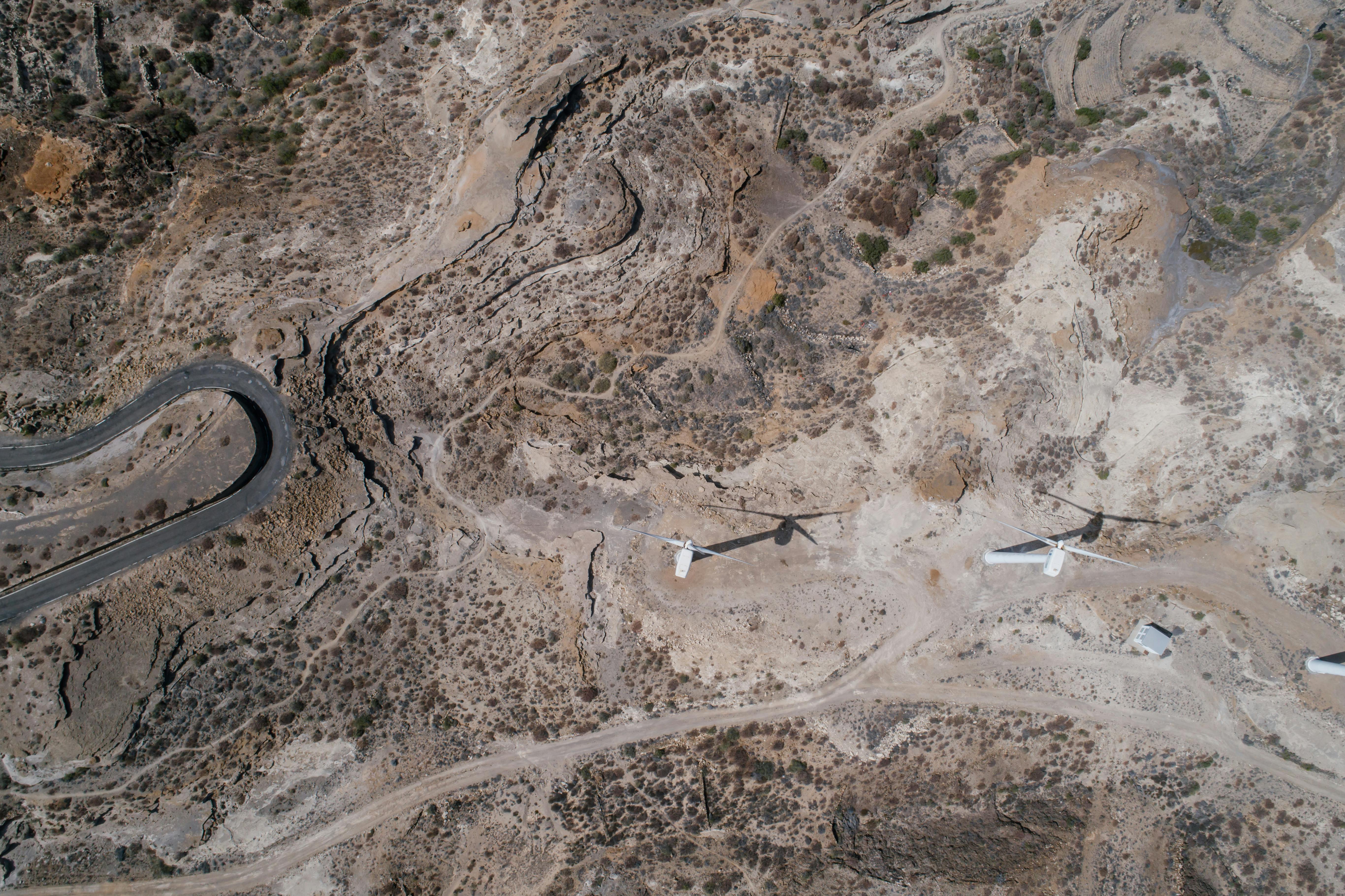 Aerial view of a rocky, arid landscape with two white wind turbines casting long shadows near dirt roads.