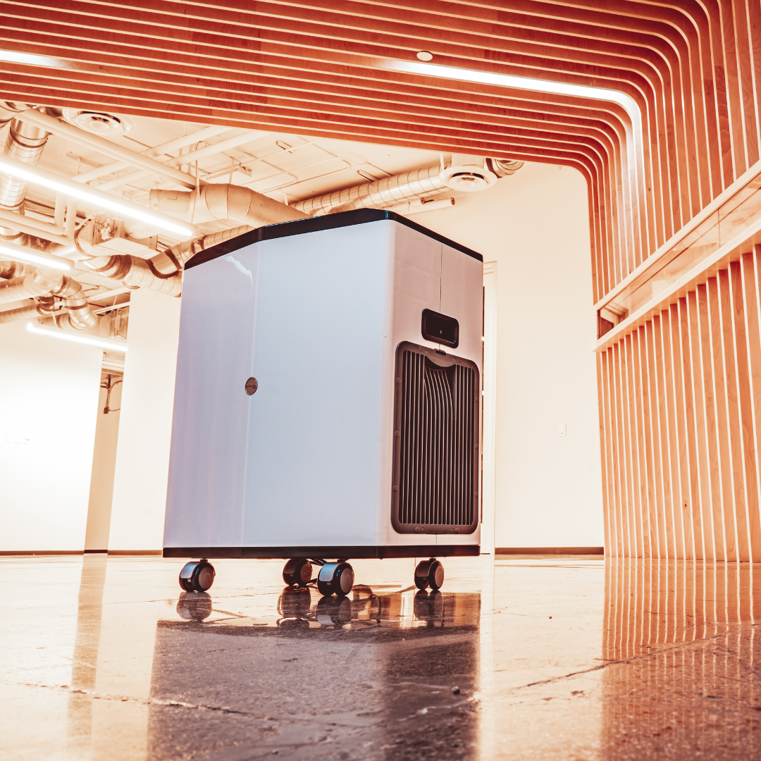 Modern white and black air purifier on wheels placed on polished floor in a room with wooden ceiling panels and exposed pipes.