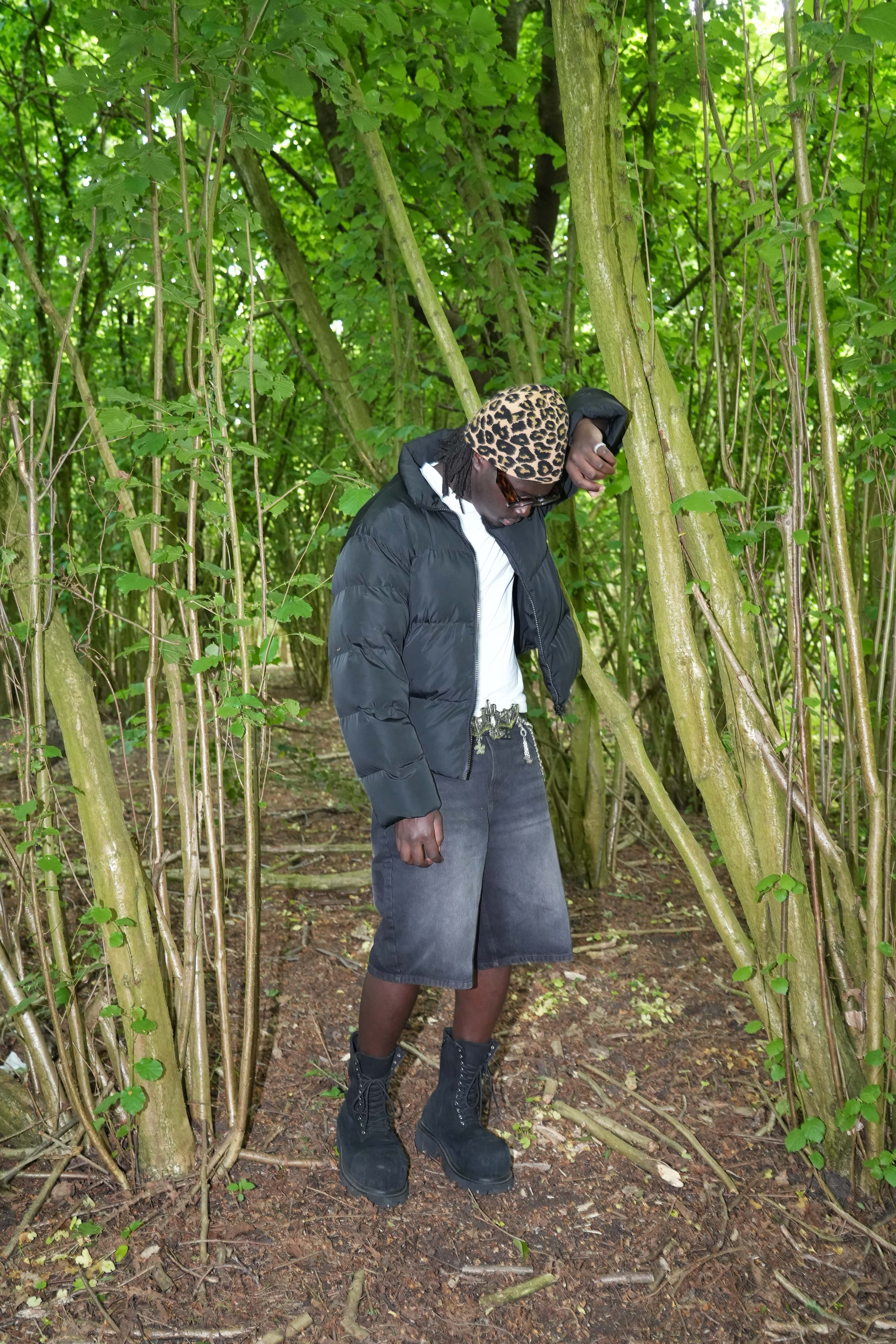 Person wearing a black puffer jacket, leopard print beanie, black shorts, white shirt, and black boots standing in a forest with dense green foliage.