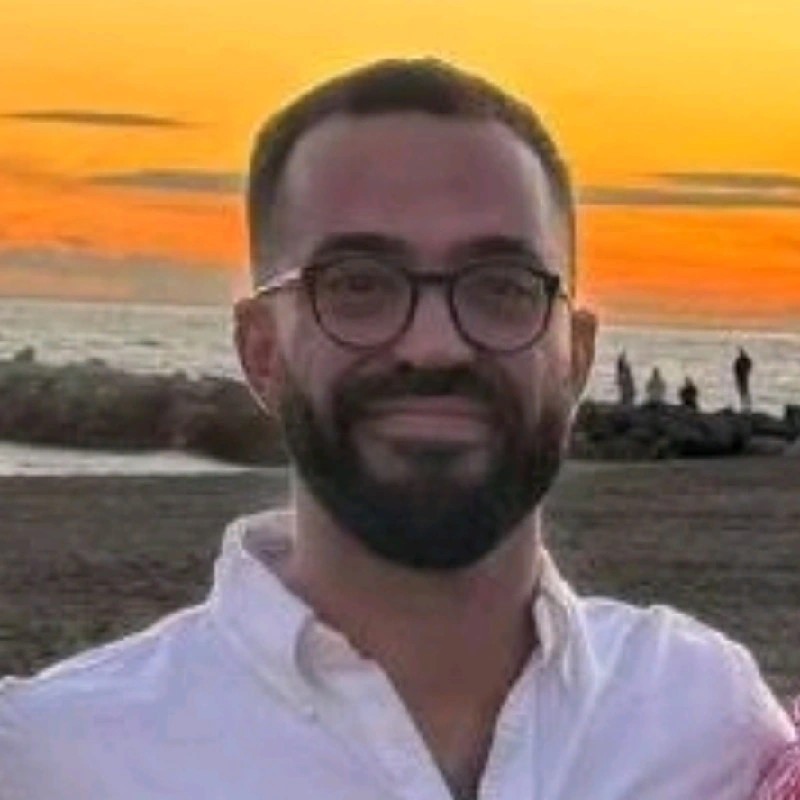 Man with glasses and beard smiling with a beach and orange sunset in the background.