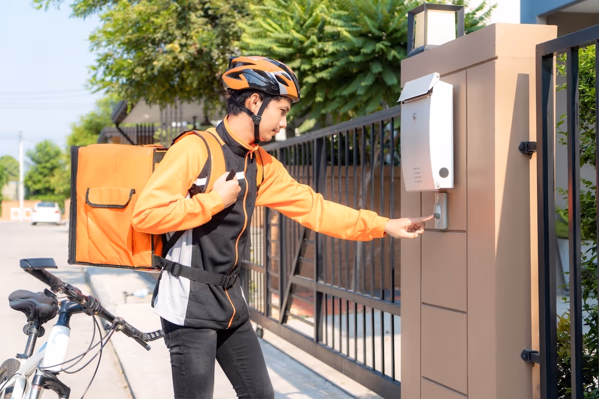 Bicycle delivery man wearing an orange uniform and helmet ringing a doorbell at a residential gate to deliver a package