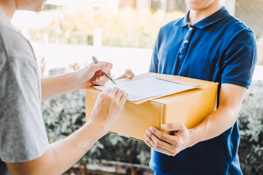 Close-up of a customer signing a delivery receipt for a cardboard package handed over by a uniformed courier at the doorstep