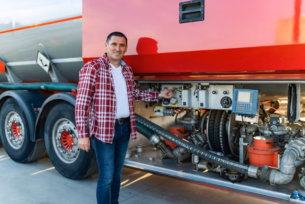 Man in plaid shirt inspecting fuel and mechanical controls on a red tanker truck, ensuring vehicle safety and maintenance compliance