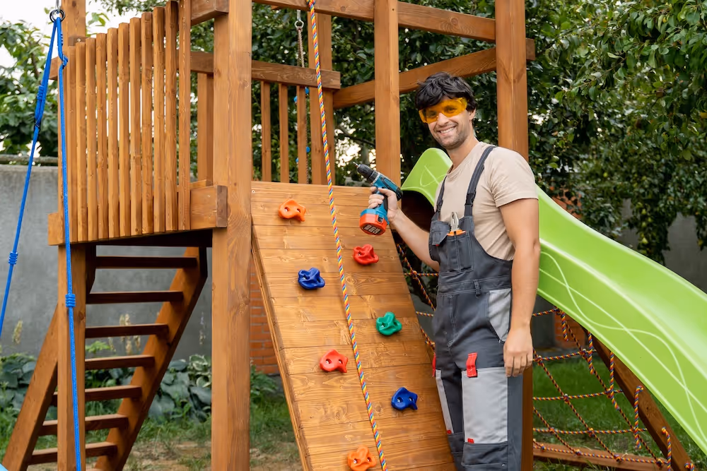 Man in protective goggles and overalls assembling a wooden backyard playset with a drill, including a climbing wall and green slide