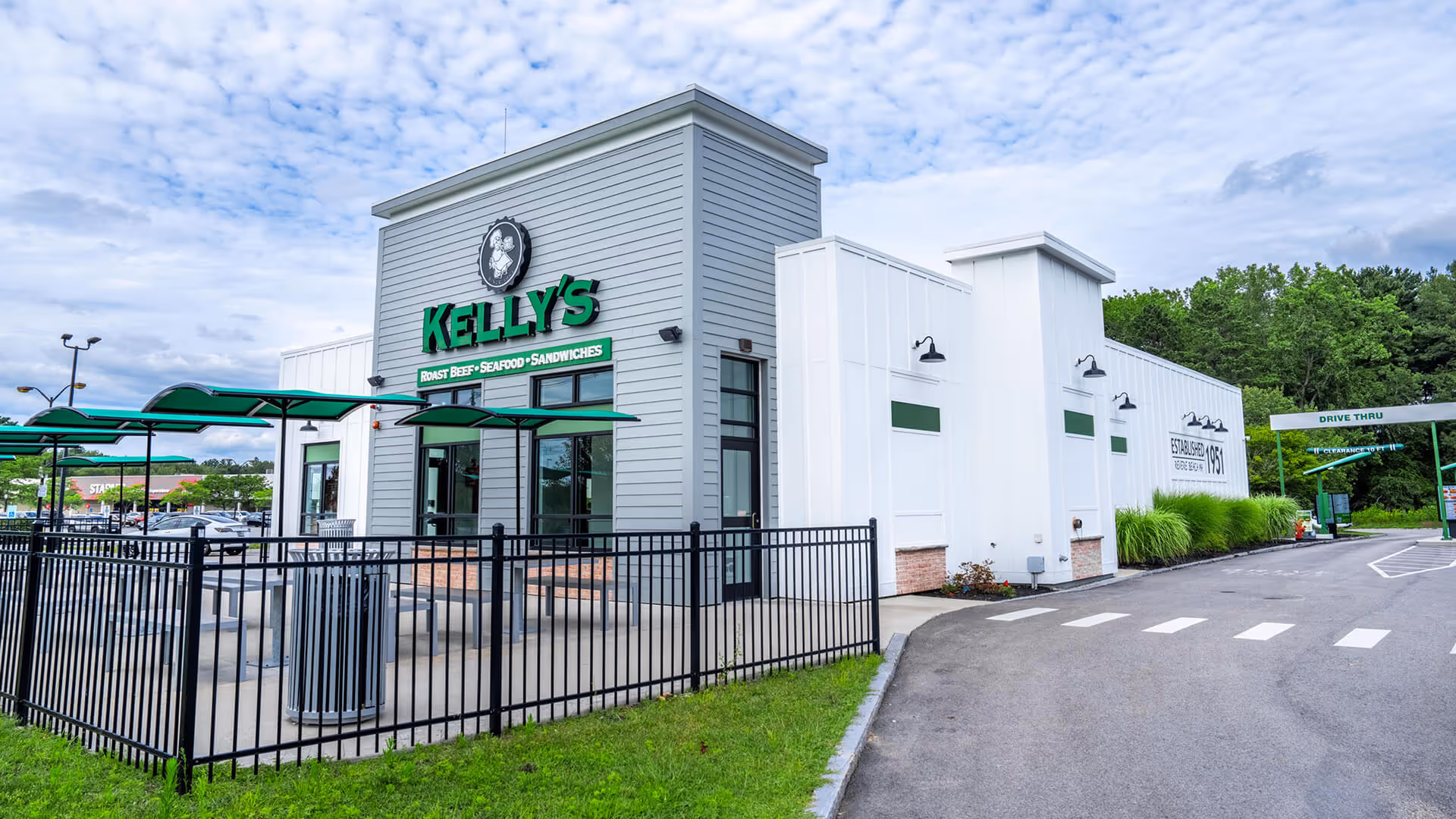 Exterior of Kelly's restaurant with green signage, outdoor seating area with green umbrellas, and a drive-thru lane.