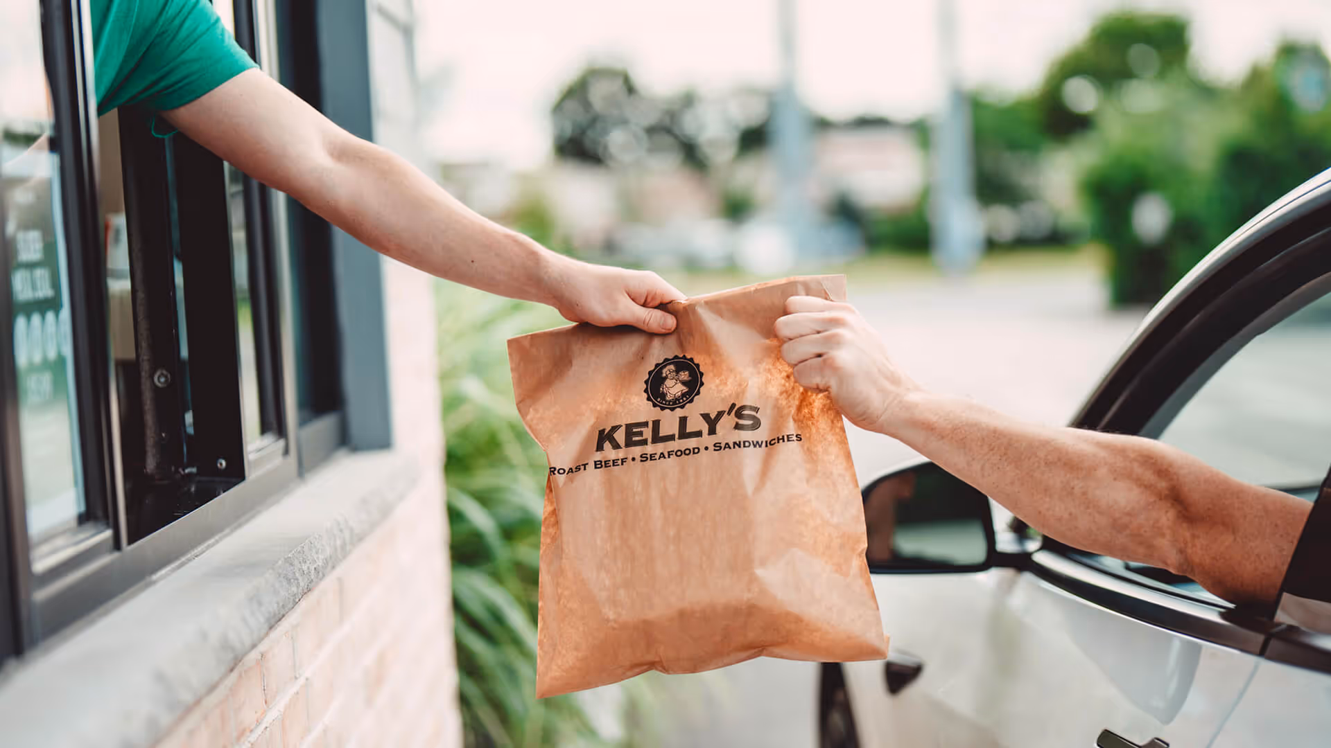 Drive-through employee handing a paper bag with Kelly's logo to a customer in a white car.