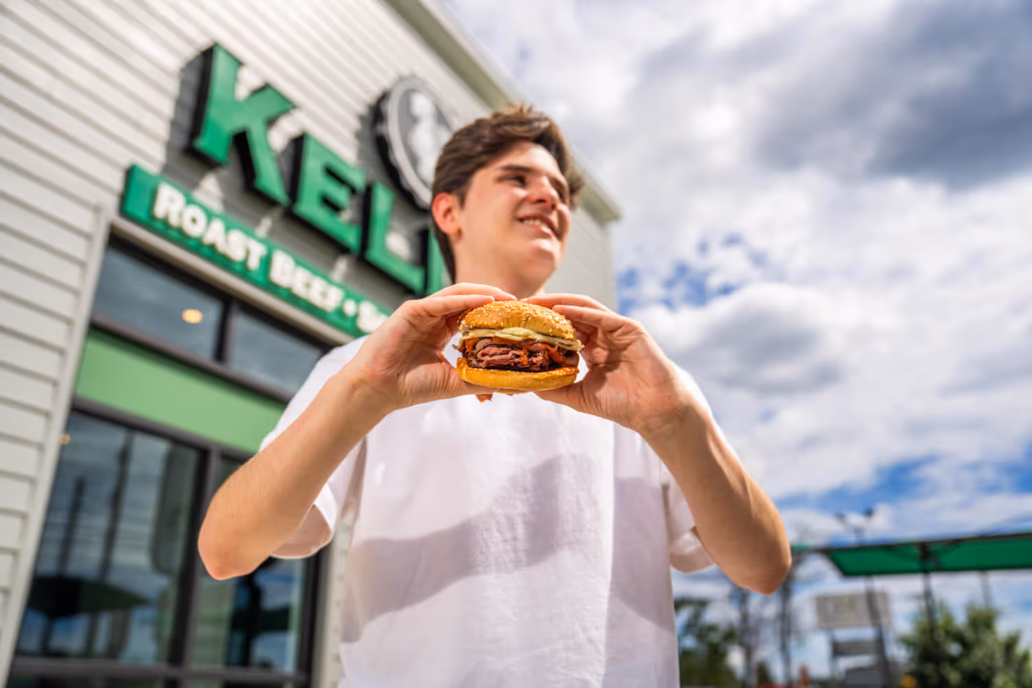 Smiling young man in a white t-shirt holding a sandwich with roast beef and bacon outside a Kelly's restaurant.