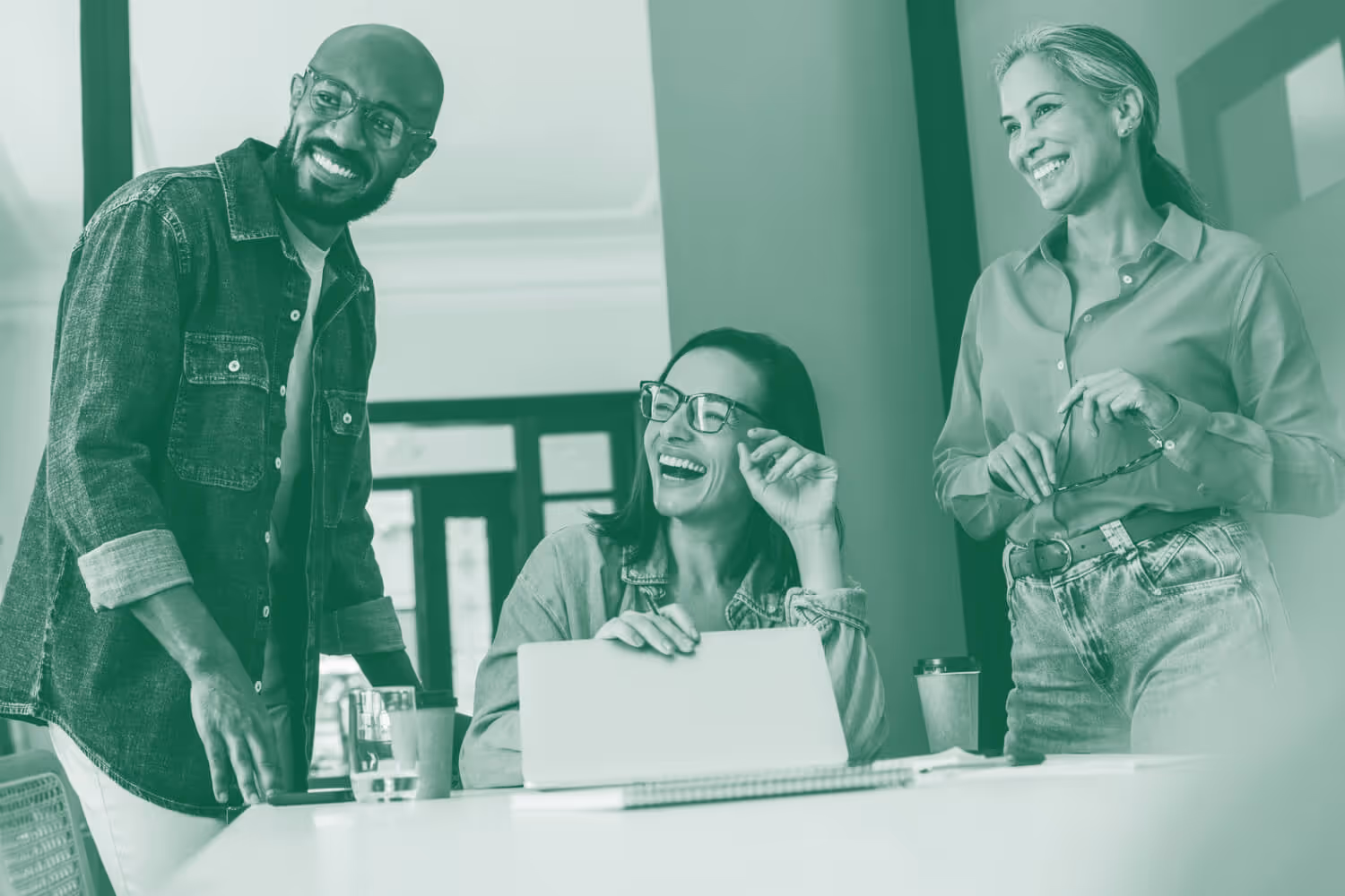 Three coworkers smiling and laughing together in a casual office setting around a table with a laptop.