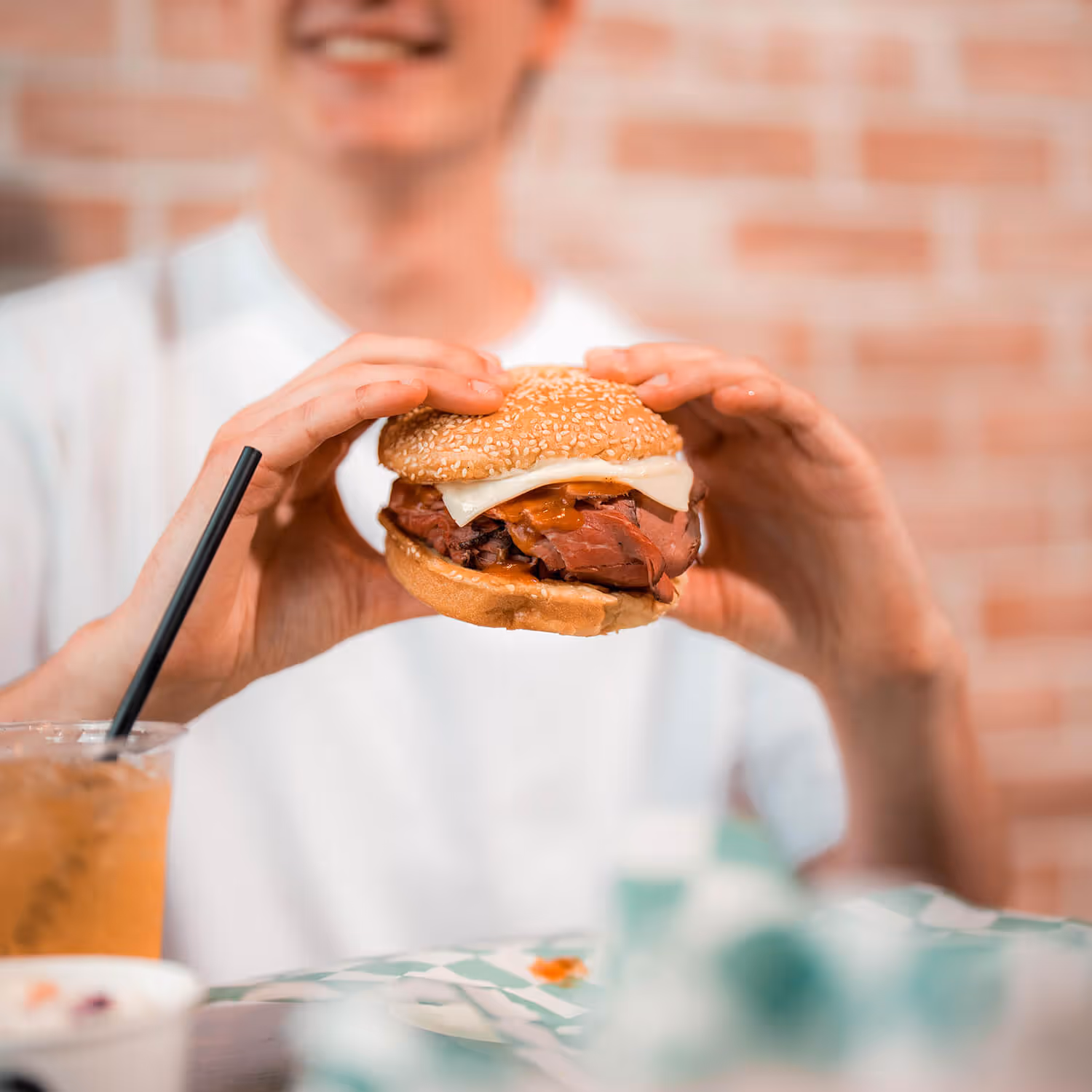 Person in white shirt holding a sandwich with sliced meat and cheese, with a drink and food container on the table.