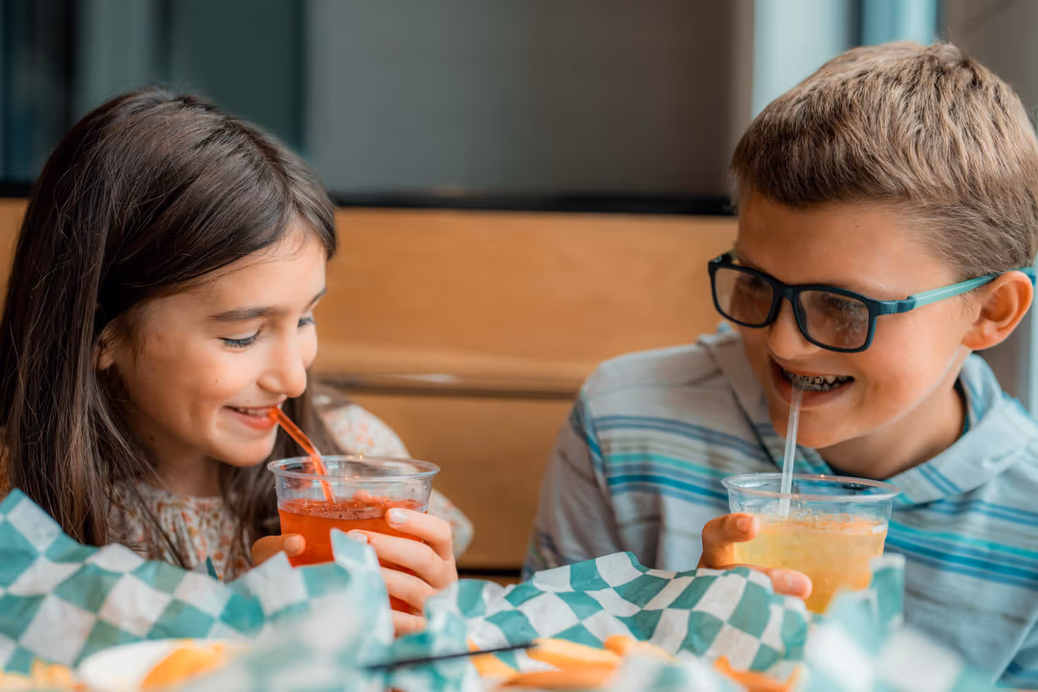 A girl and a boy smiling and drinking beverages with straws at a table with checkered paper.