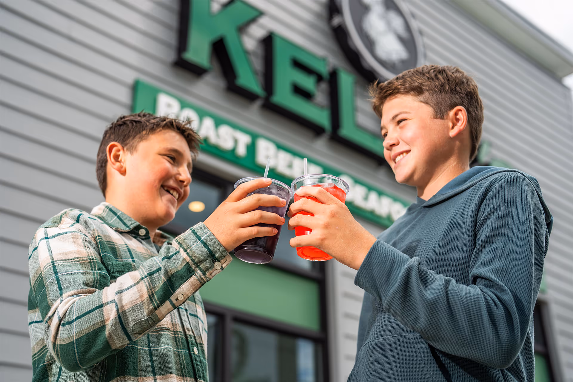 Two smiling boys clinking plastic cups with colorful drinks outside a building with a sign that reads 'ROAST BEANS'.