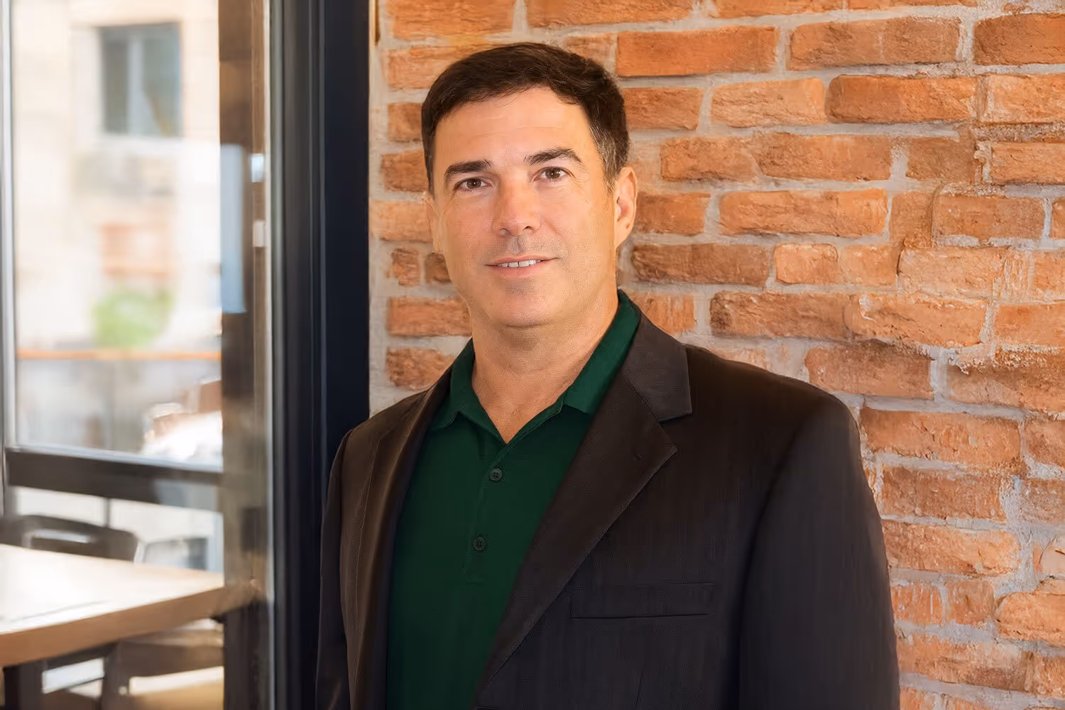 Man in dark blazer and green polo shirt standing in front of a brick wall near a window.