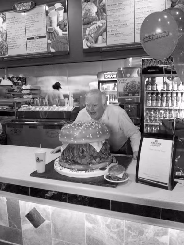 Man behind counter posing with an oversized hamburger display in a restaurant.