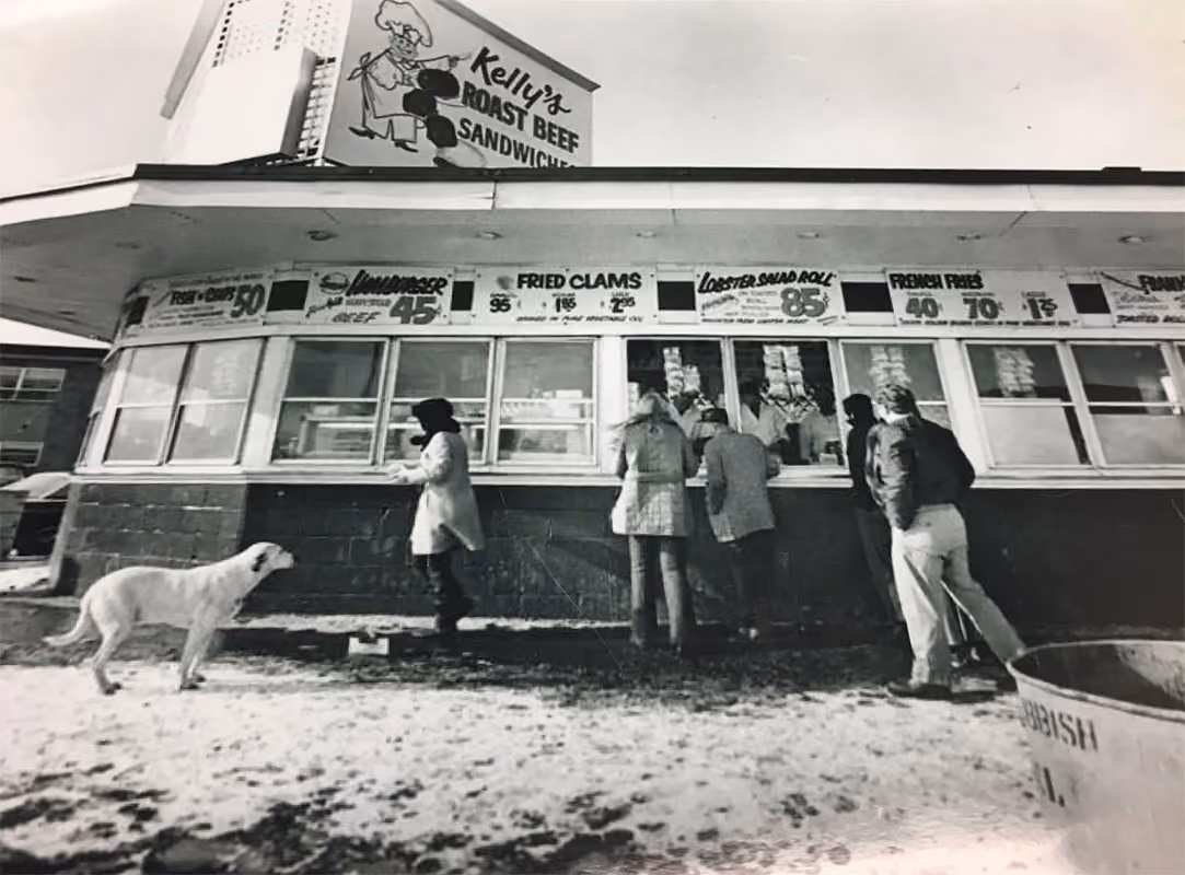 Black and white photo of people standing at Kelly’s Roast Beef counter with a dog nearby outdoors.