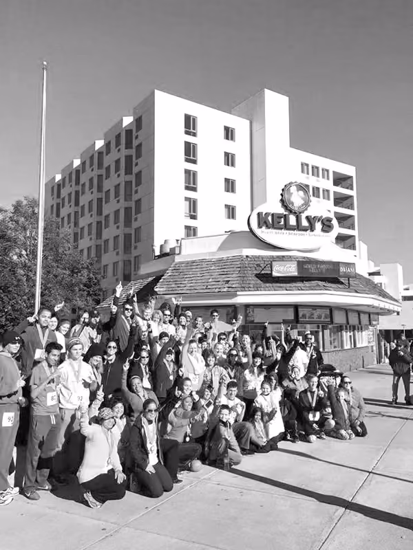 Large group of people posing happily outside Kelly's restaurant on a sunny day.