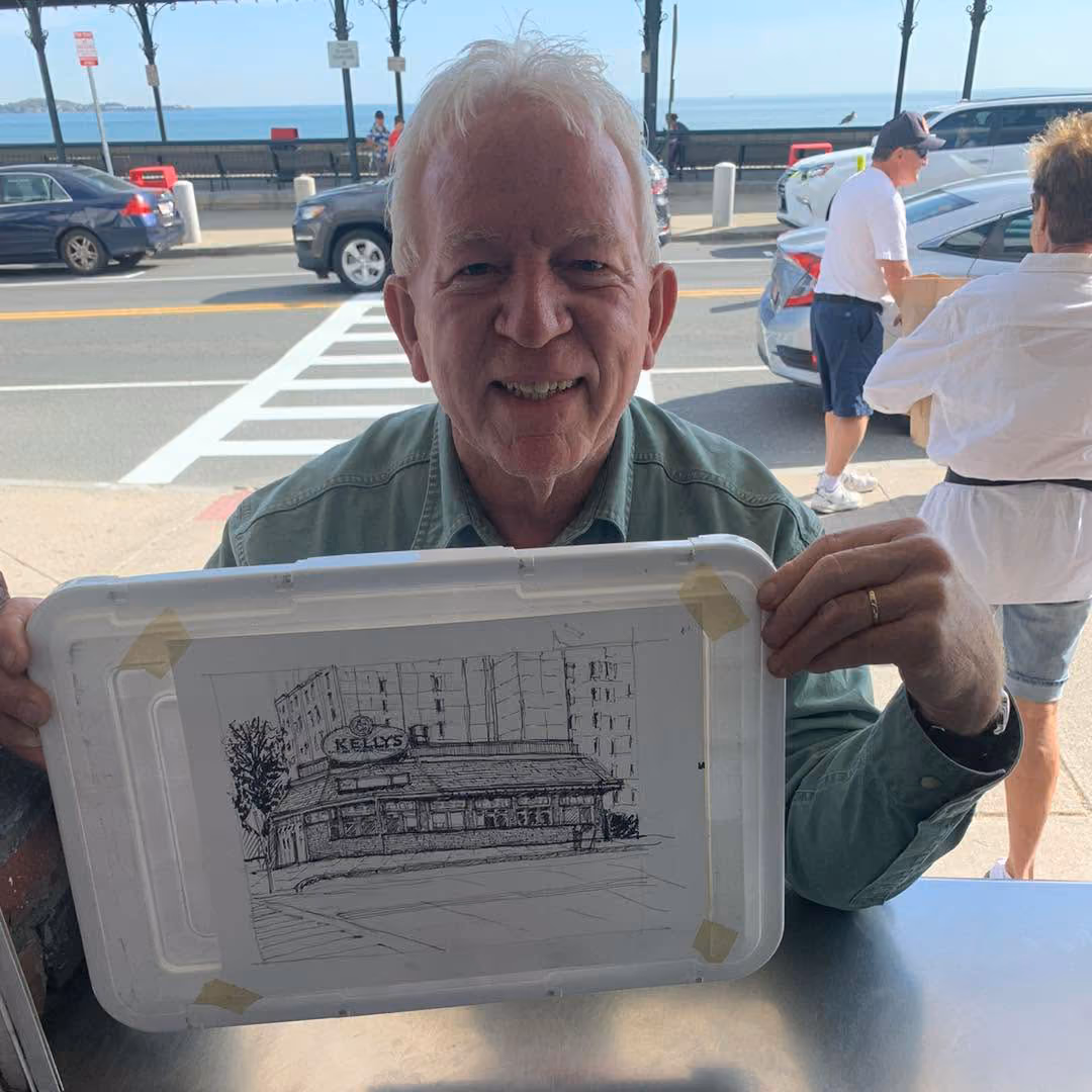 Older man with white hair holding a sketch of a building labeled Kelly's near a street and cars.