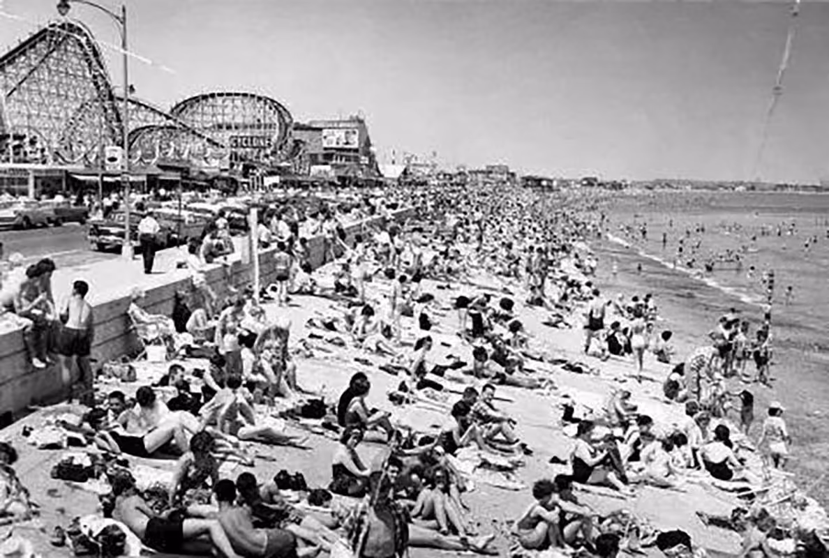 Crowded beach with people sunbathing, roller coaster, and amusement park rides in the background.