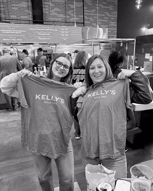 Two smiling women holding up Kelly's Since 1951 t-shirts inside a restaurant.