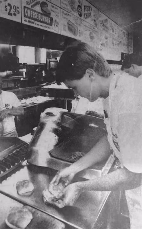 Young person preparing hamburger buns at a kitchen counter with menu signs overhead.