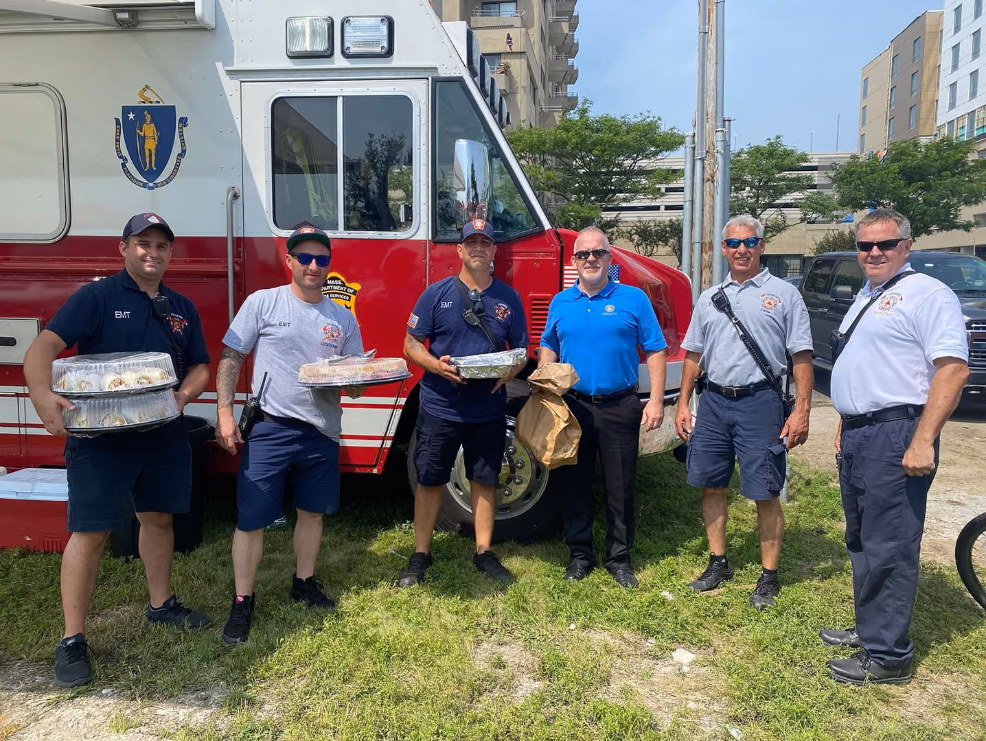 Group of six fire and emergency workers standing in front of a fire truck holding food trays and bags.