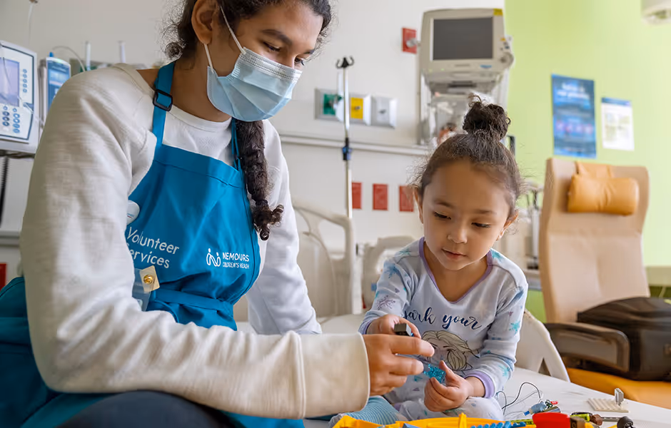 Masked female volunteer in a blue apron helps a young girl with a toy in a hospital room.