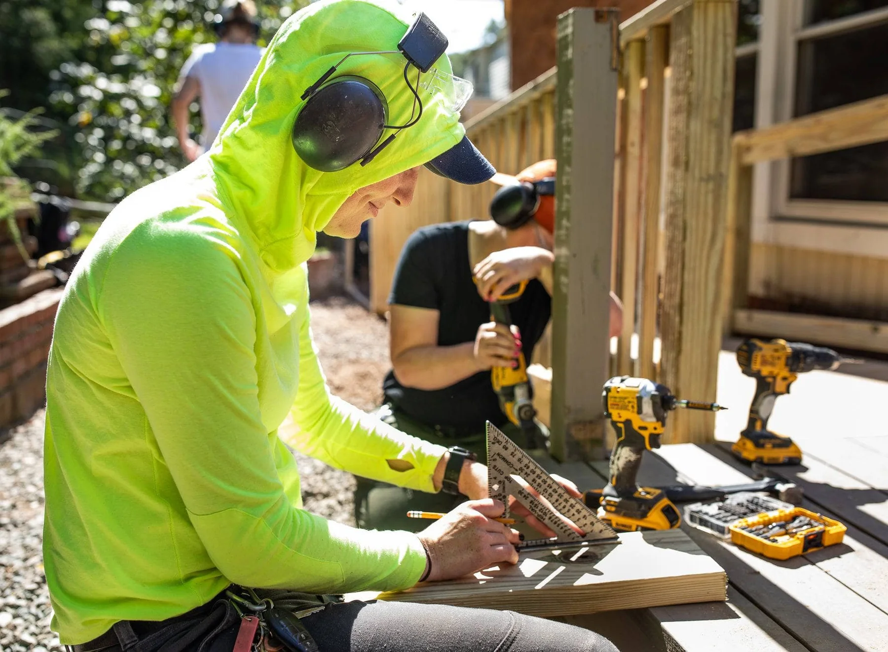 Two Hope Renovations workers with ear protection using a square ruler and power drills while working on wooden boards outdoors.
