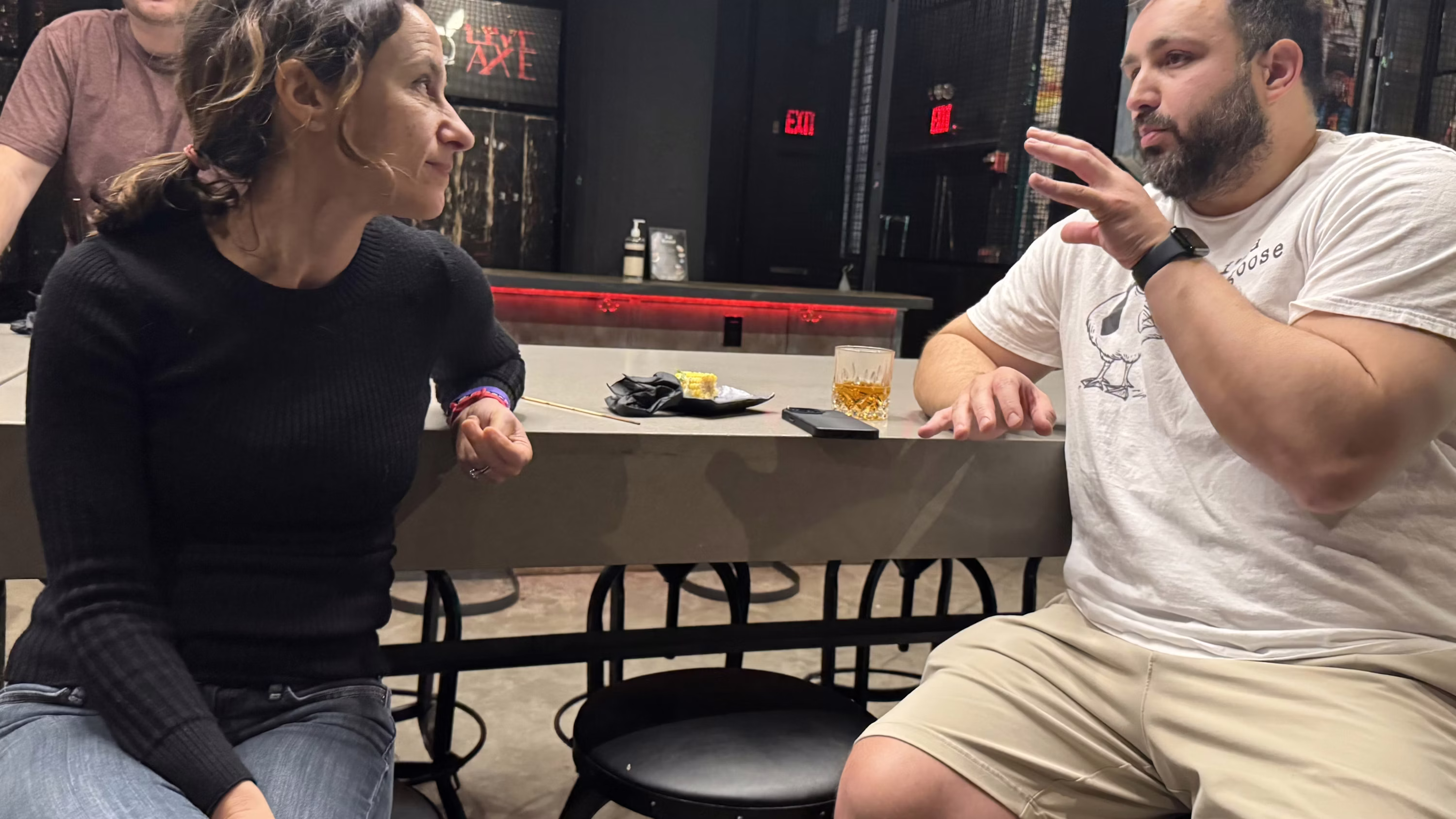 A man and woman sitting at a table in a dimly lit bar having a conversation, with a drink and snacks on the table.