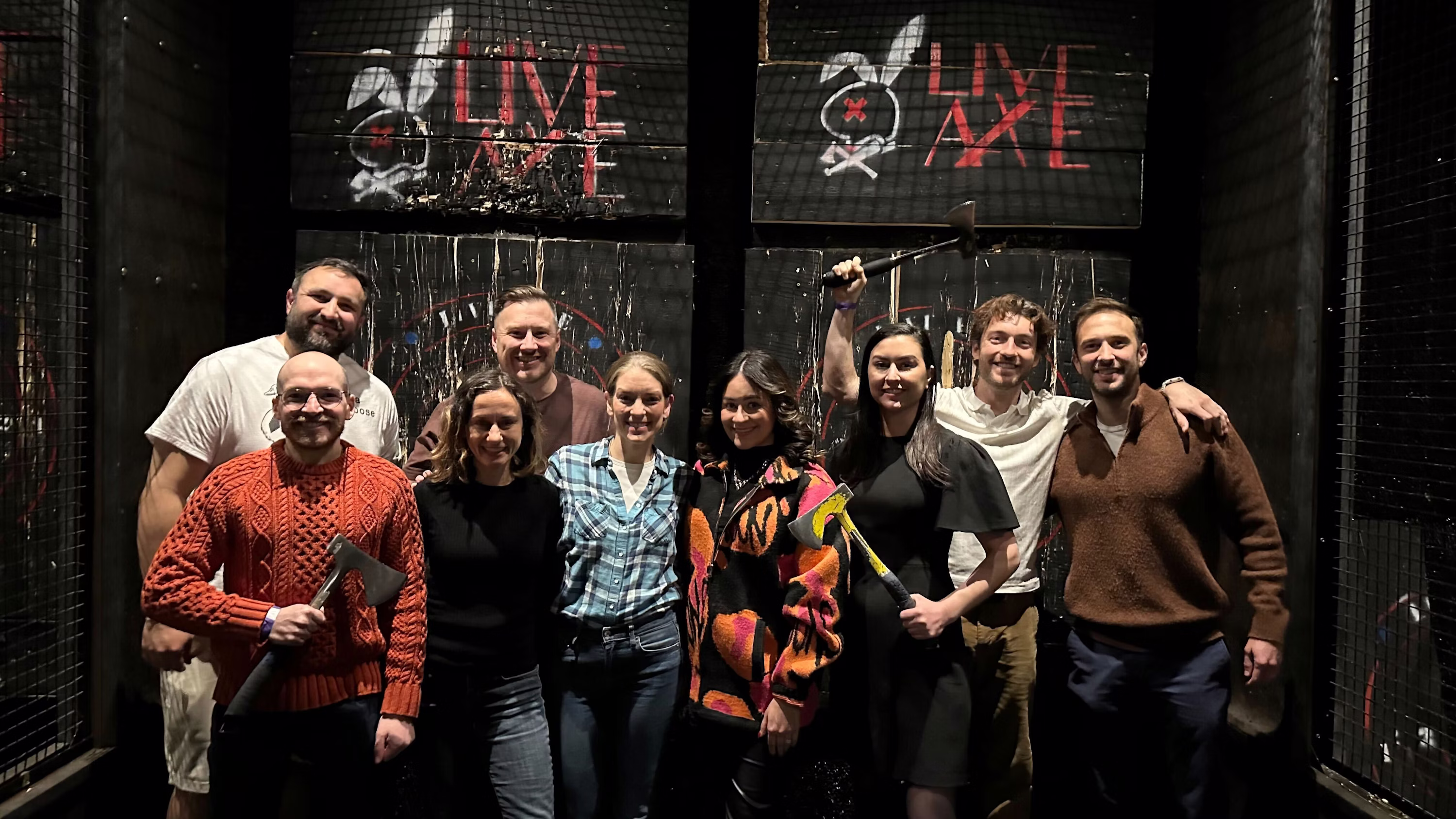 Group of eight people standing inside an axe throwing venue with axes, smiling at the camera in front of wooden targets with 'LIVE AXE' signs.