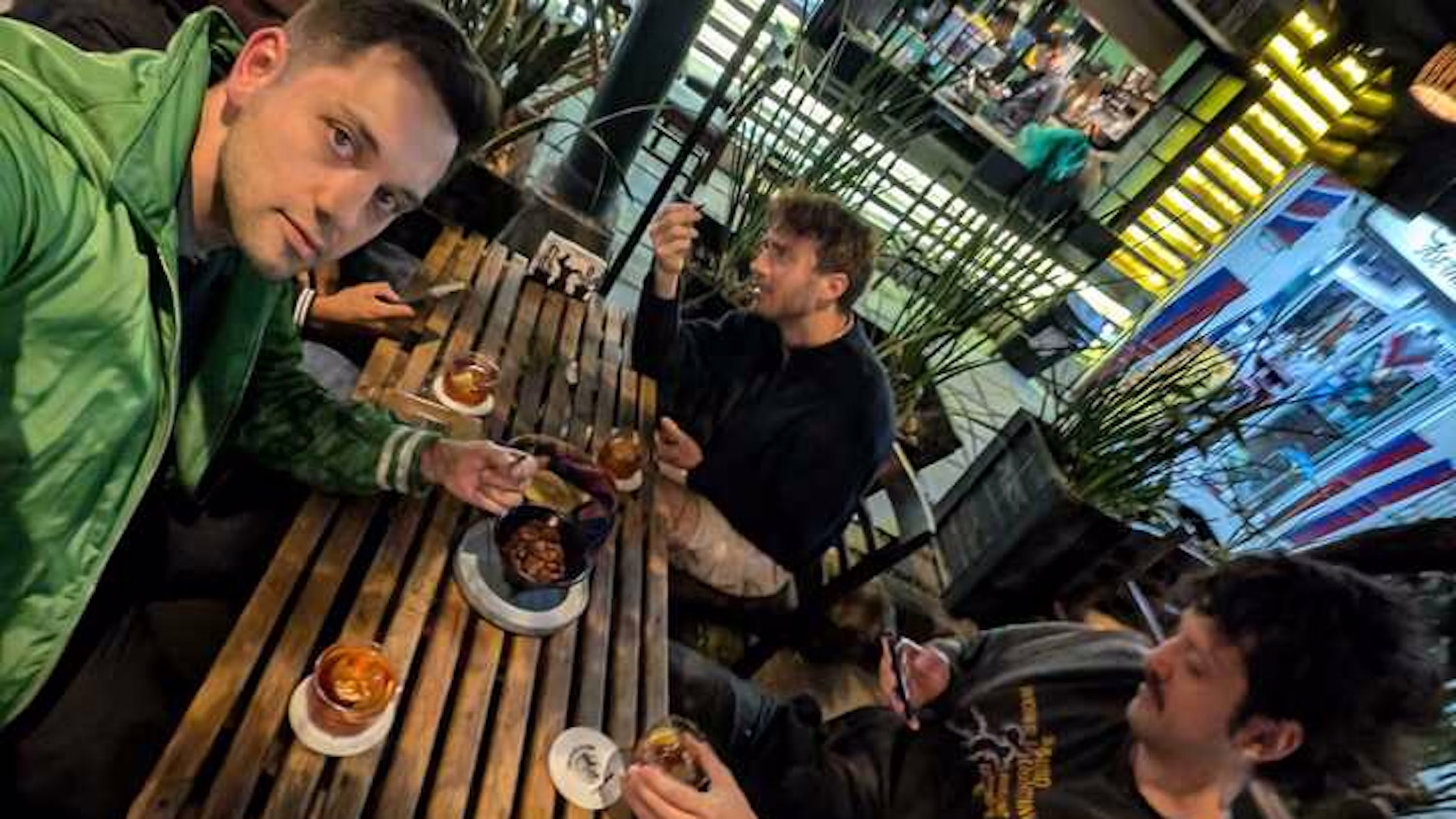 Three men sitting at a wooden table outdoors, enjoying drinks and snacks in a lively, modern setting with plants and bright lighting.